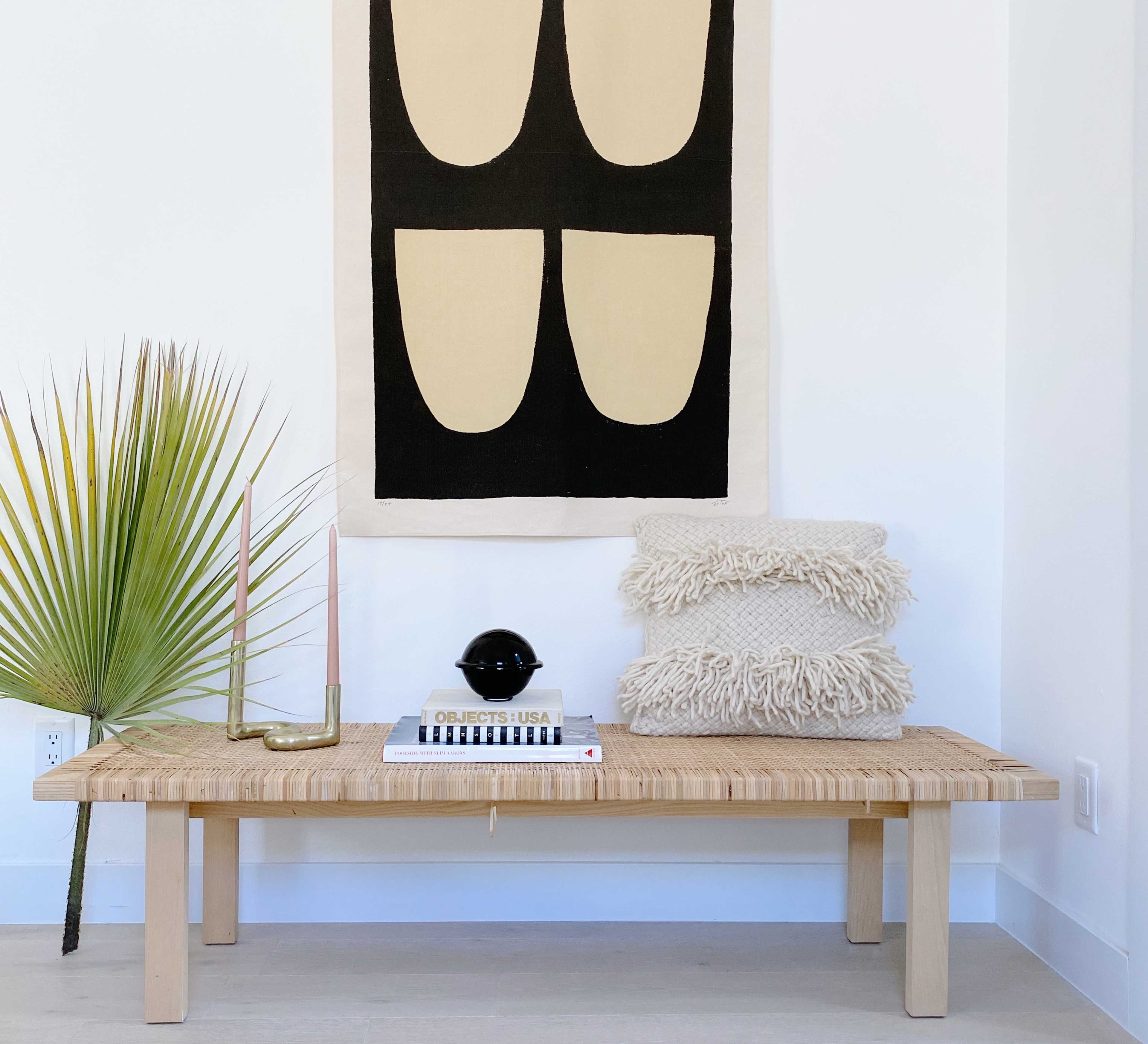 A wooden bench with a woven top is positioned against a white wall, featuring a large abstract wall hanging, a potted palm leaf, a candle holder with candles, a small black lamp, and neatly stacked textured pillows.