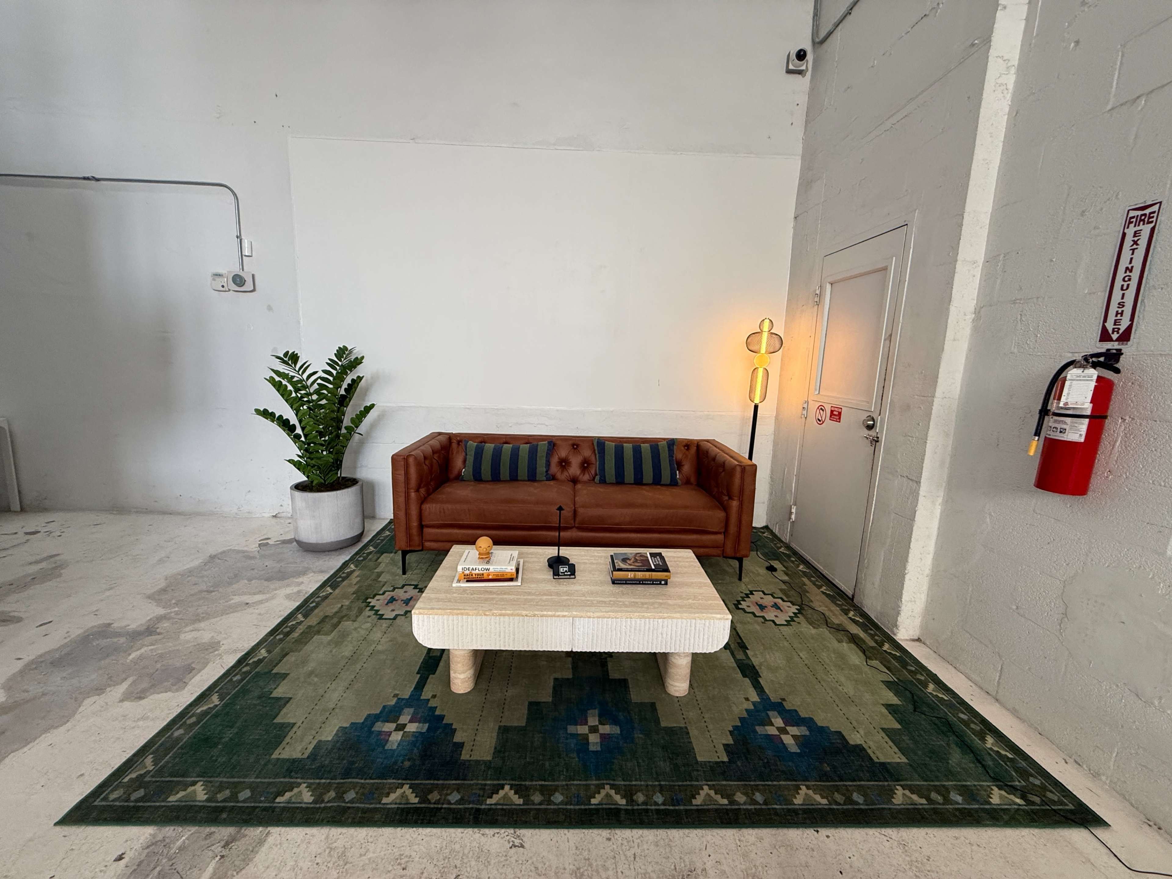 The image shows a minimalist seating area with a brown leather couch, a white coffee table, and a large green rug, accompanied by a potted plant and wall-mounted fire extinguisher in a well-lit, neutral-colored room.