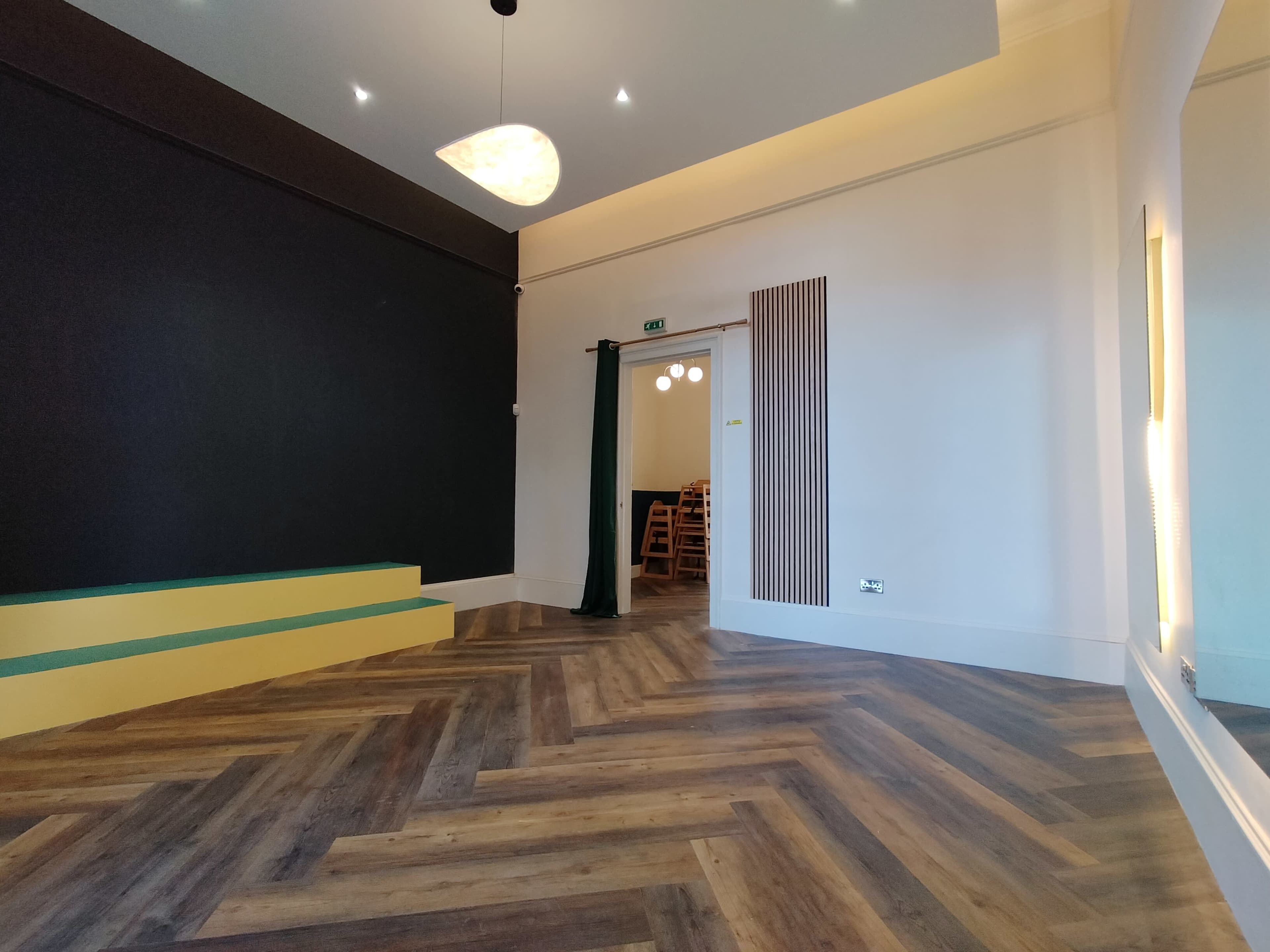 A spacious interior room with dark walls, a wooden chevron floor, a ceiling light fixture, and a doorway leading to a dining area with wooden chairs visible.