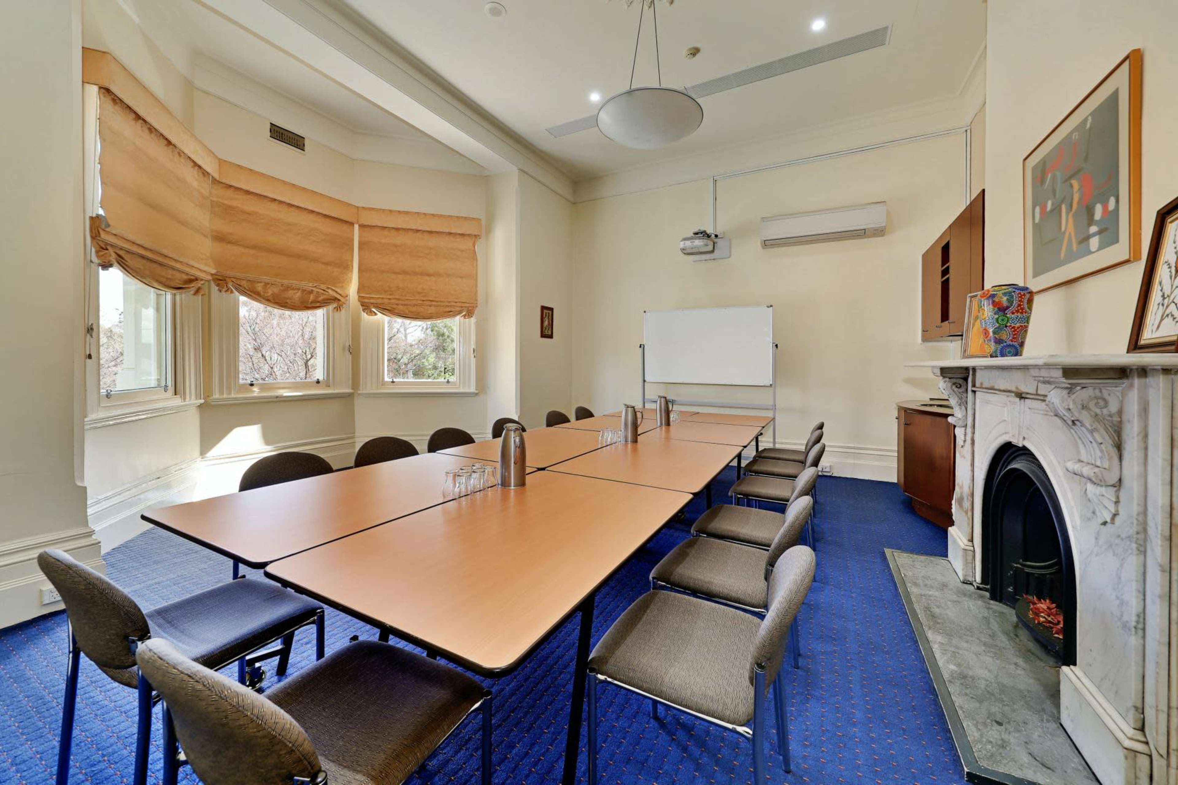 A rectangular meeting room features a long table with several chairs, large windows with Roman shades, and a whiteboard on the wall.