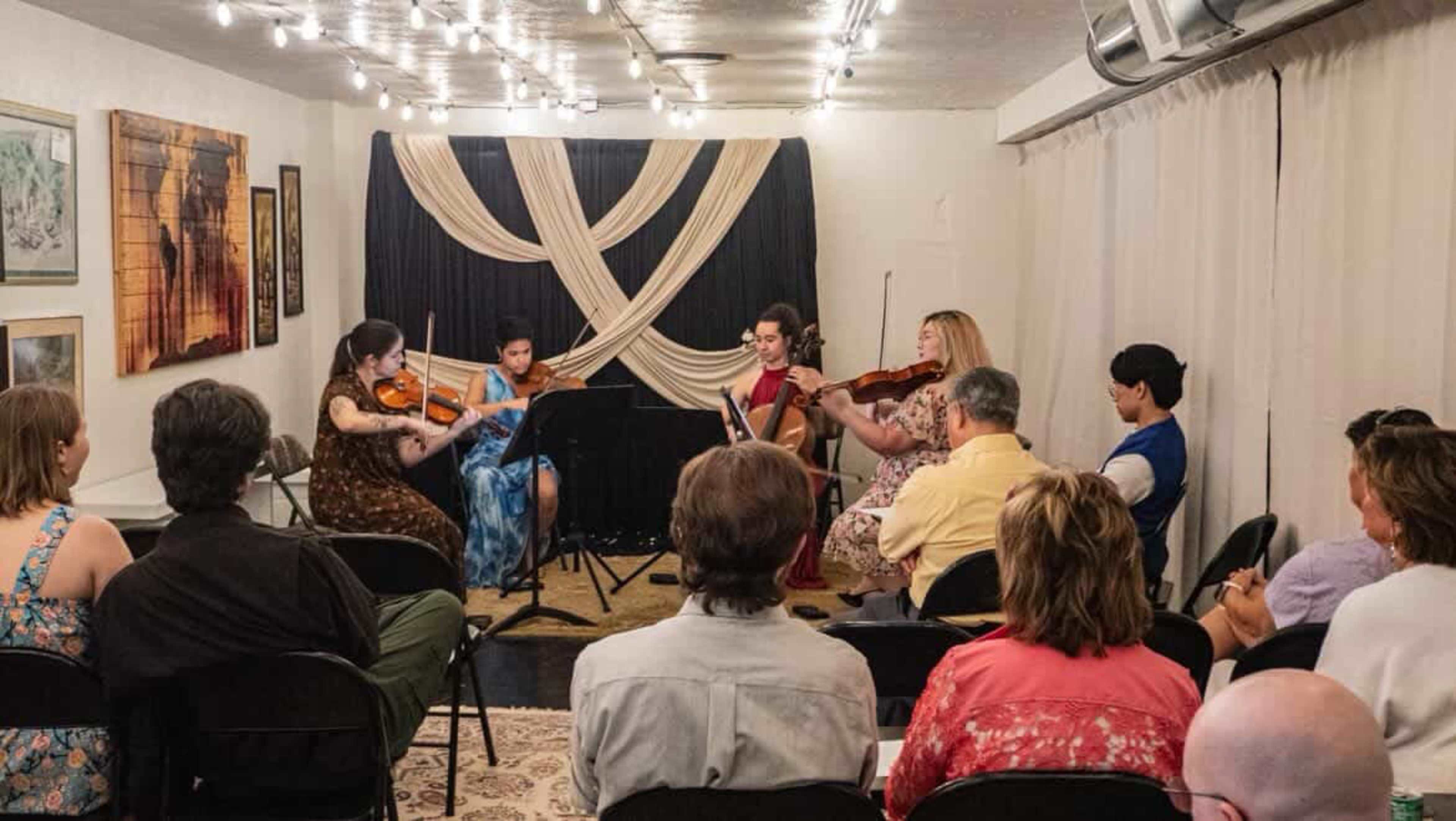 A small group of musicians performs with string instruments in an intimate setting, while an audience observes seated on folding chairs.
