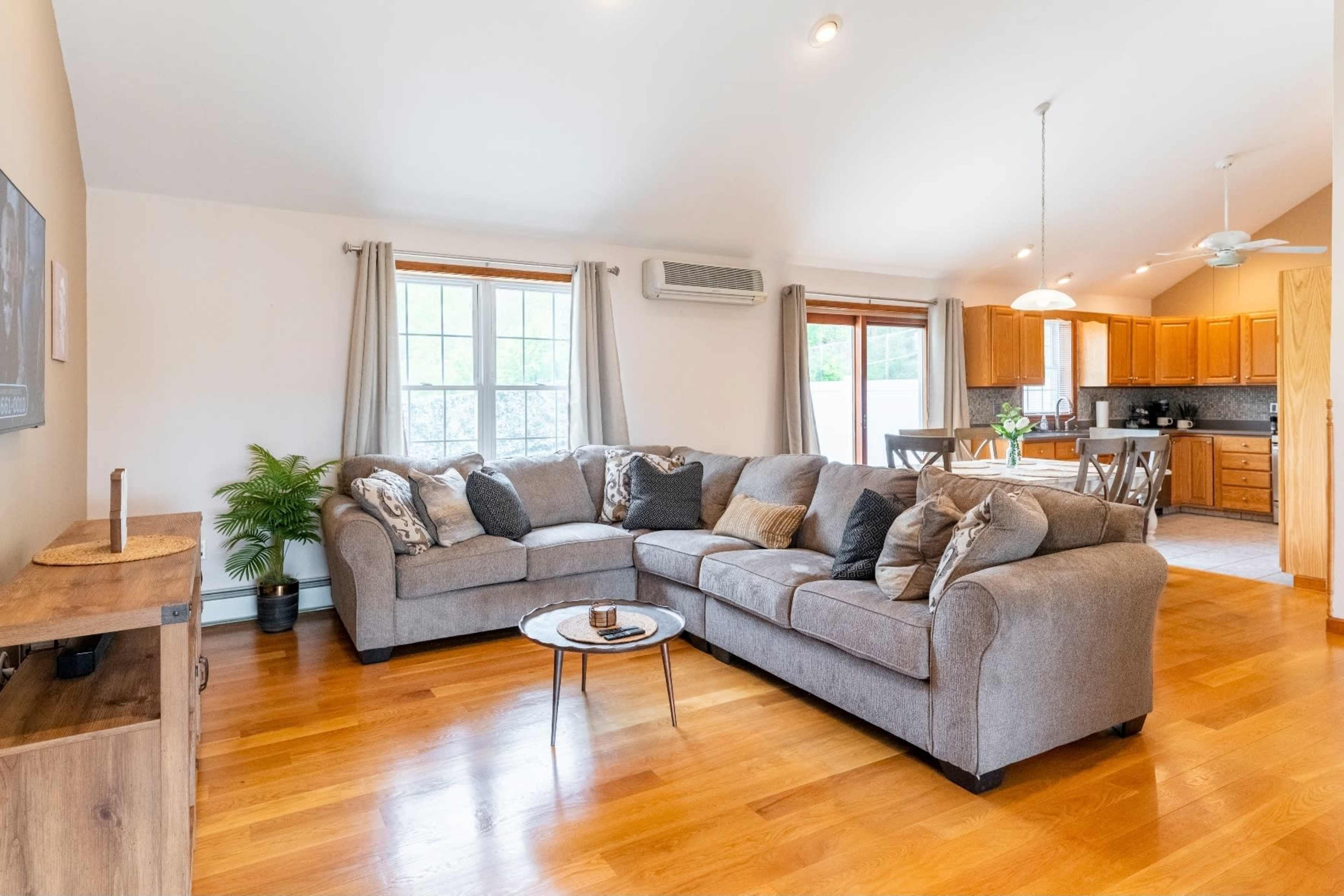 A spacious living room features a large gray sectional sofa, wooden floors, and an open layout leading to a kitchen with wooden cabinetry.