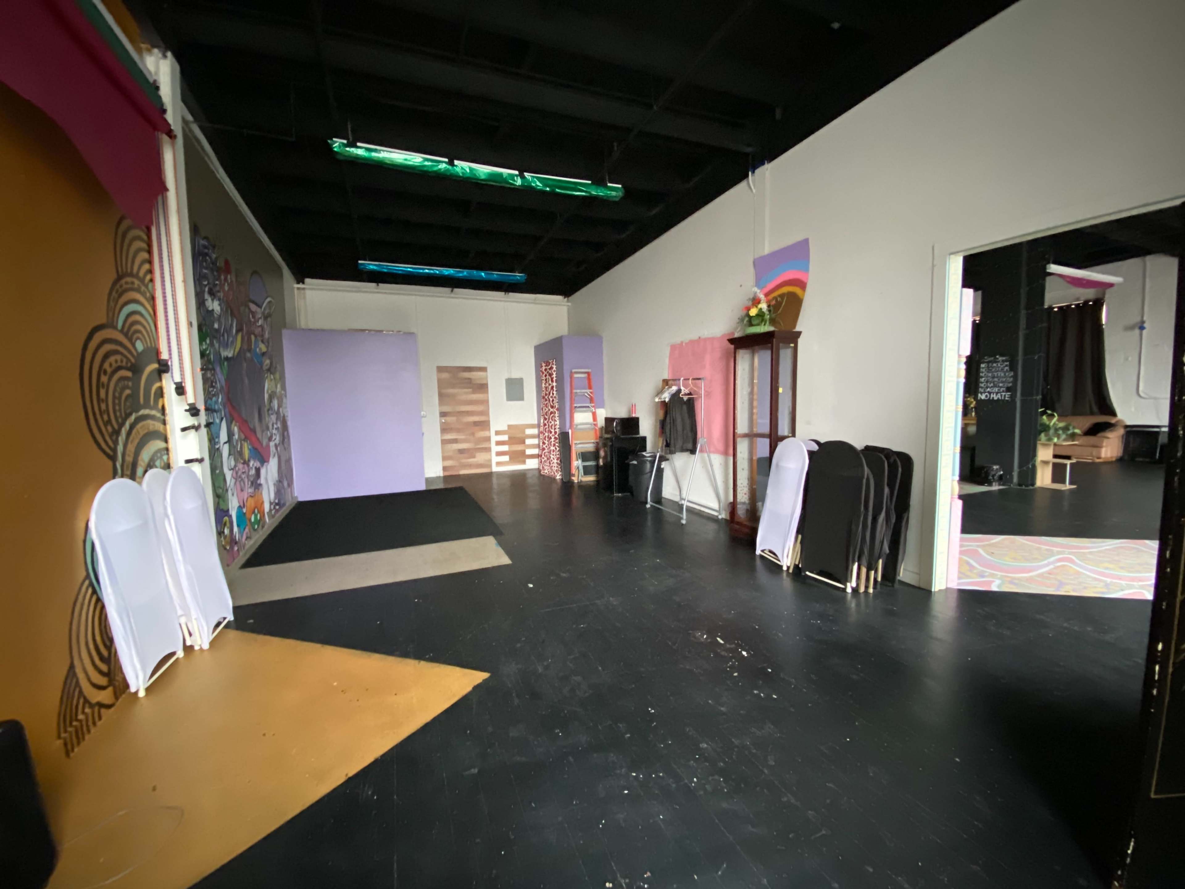 The image shows an empty studio space with black floors, colorful wall art, and several folding chairs stacked against one wall.
