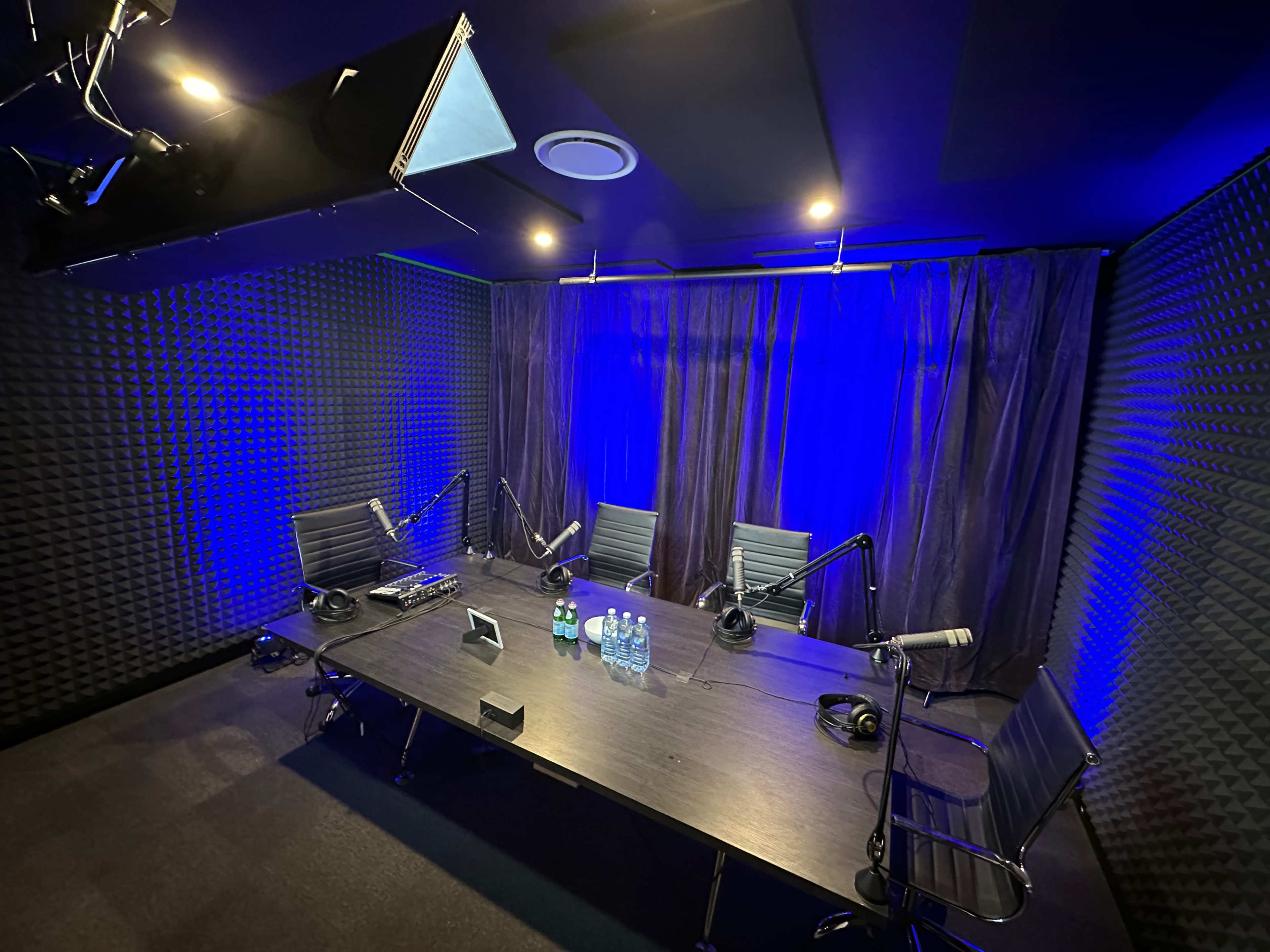The image shows a recording studio with a large table, four chairs, and multiple microphones, surrounded by soundproof walls and blue lighting.