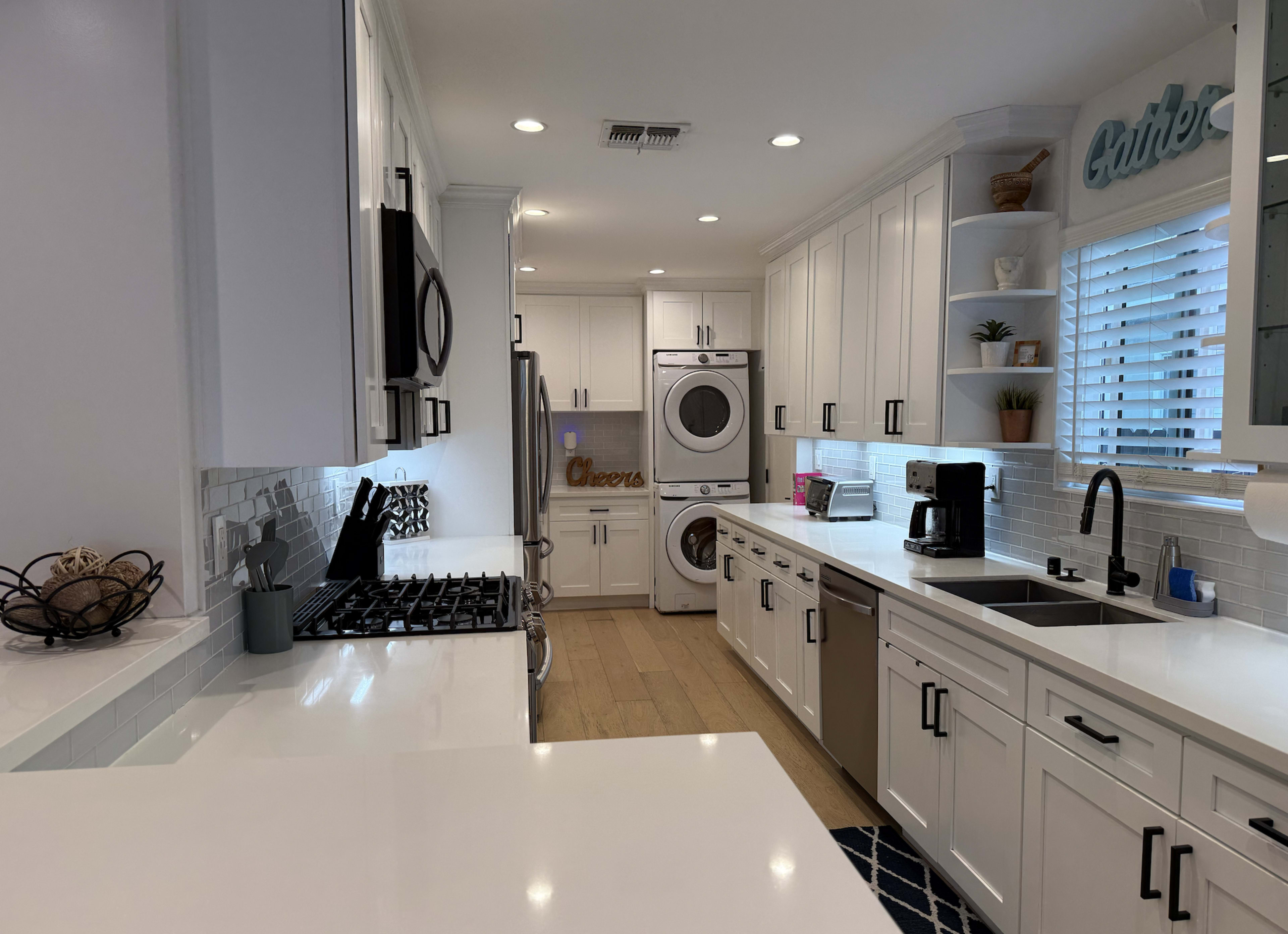 The image shows a modern kitchen with white cabinetry, stainless steel appliances, and a laundry area at the end featuring a stacked washer and dryer.
