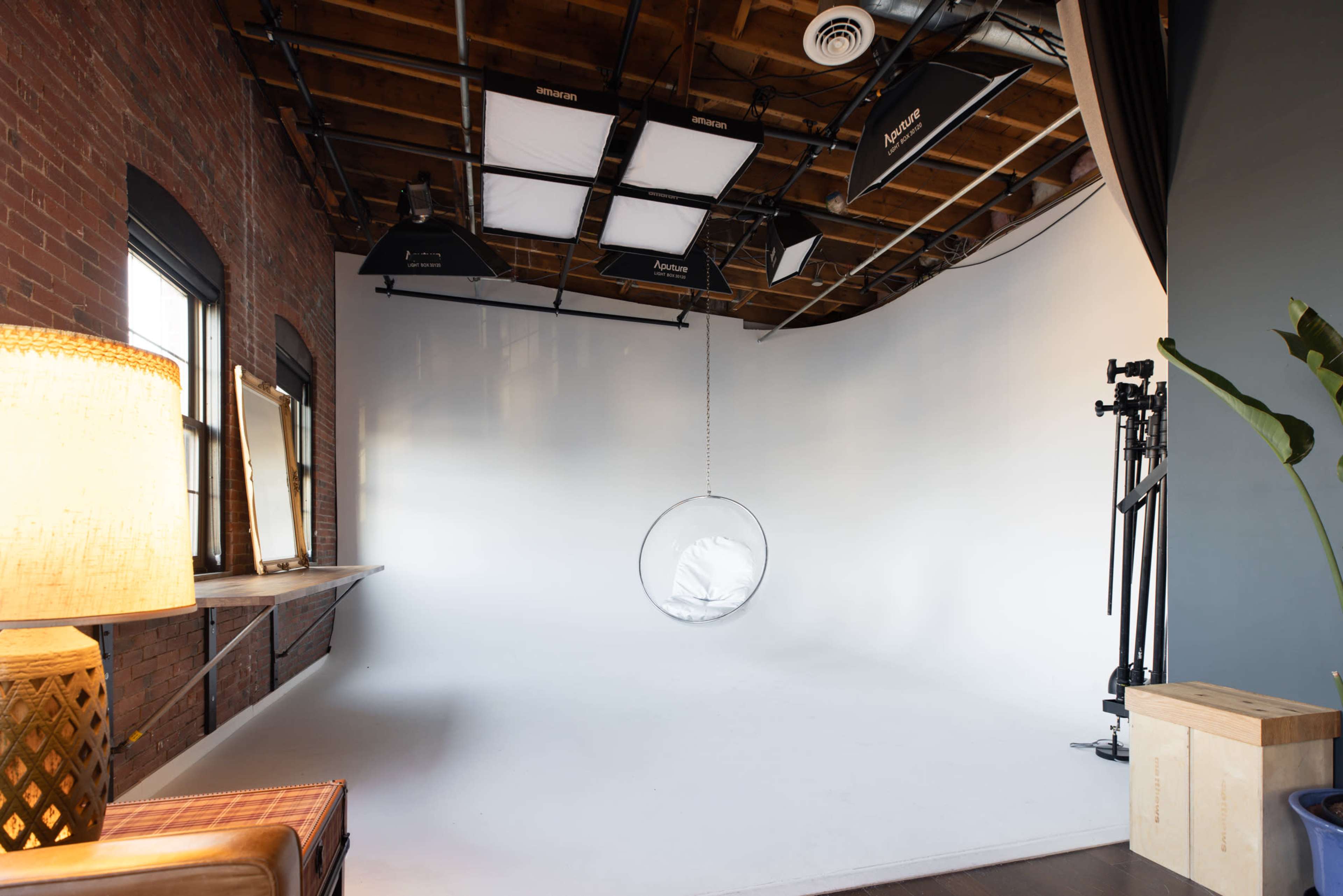 The image shows a photography studio with a white seamless backdrop, a hanging chair, lighting equipment, and a wooden table with mirrors.