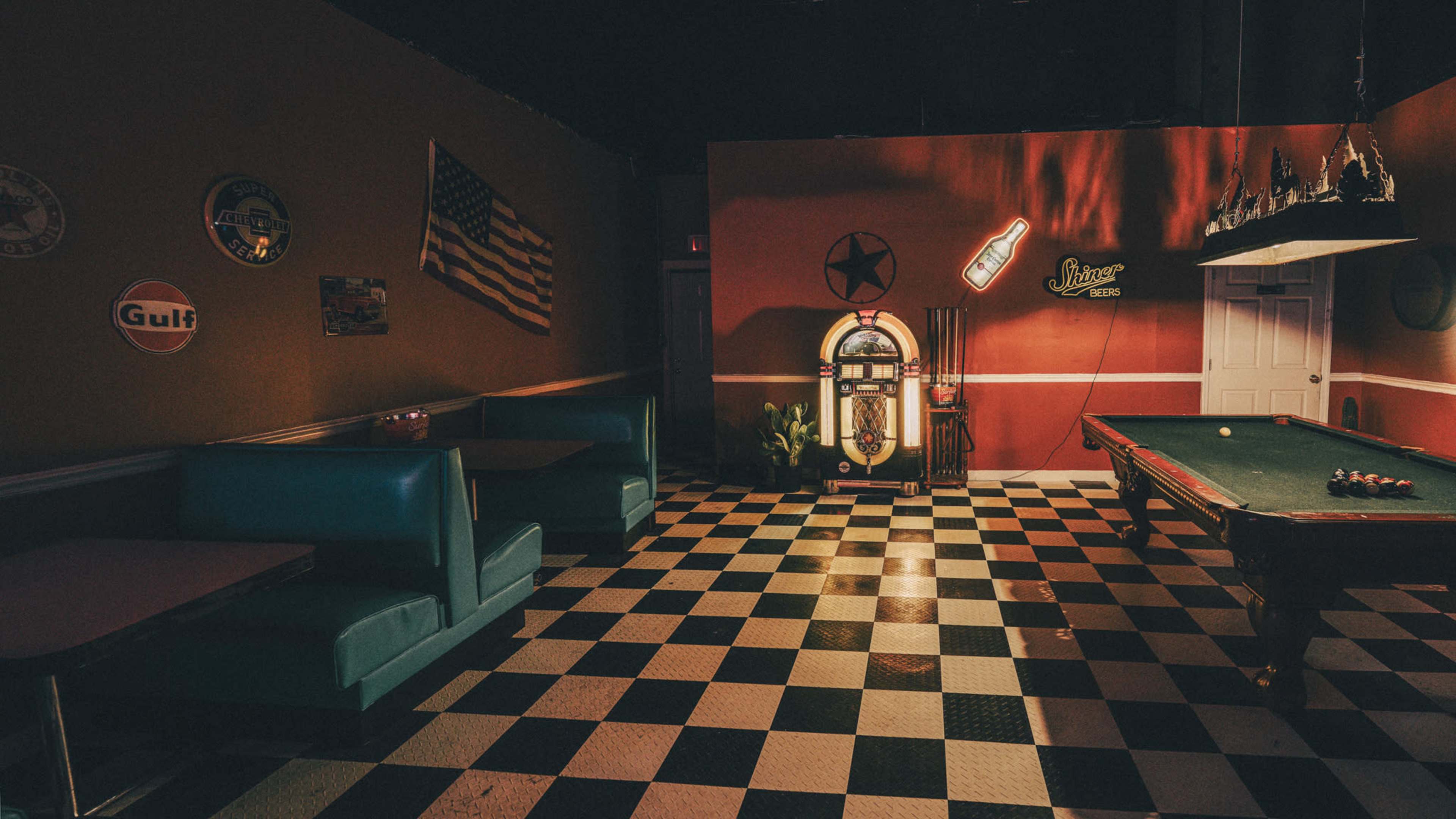 The image shows a dimly lit vintage diner with checkered flooring, featuring a jukebox, a pool table, and teal booths along the walls decorated with Americana memorabilia.