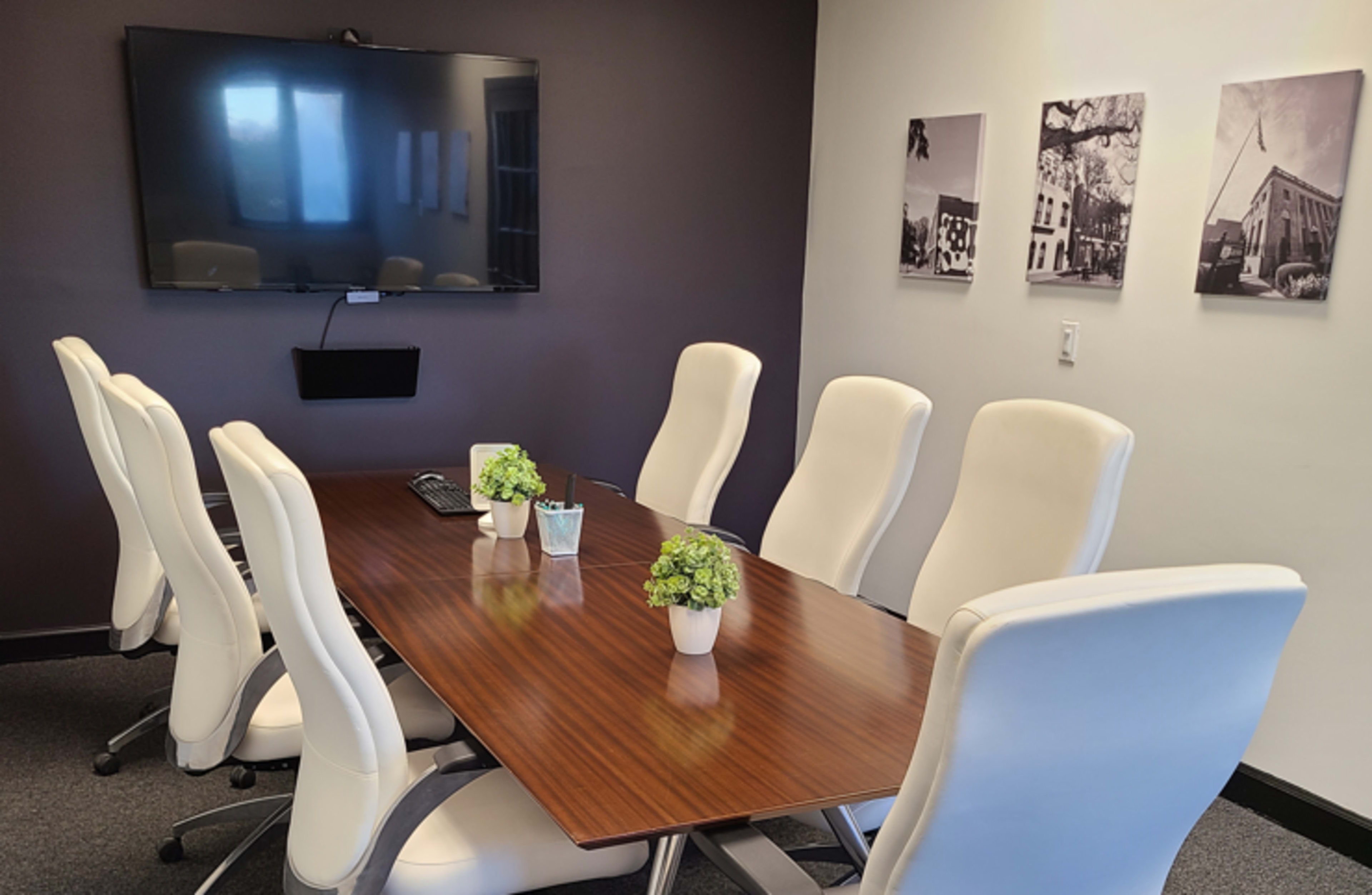 A conference room features a long wooden table surrounded by white chairs, with a large screen on one wall and framed black-and-white photographs on another.