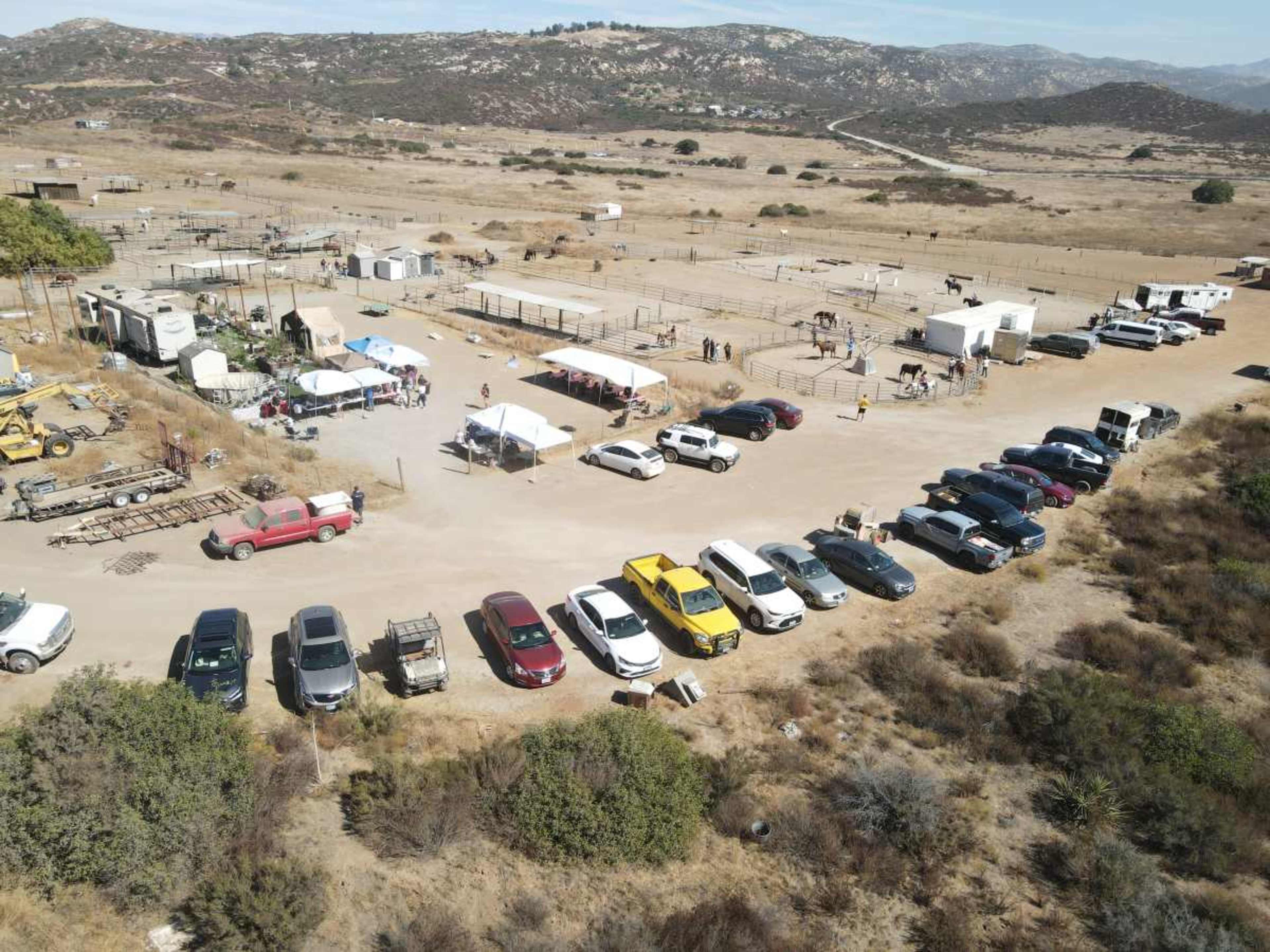 The image shows a dirt parking area filled with various vehicles adjacent to a rural site featuring tents, horse enclosures, and mountainous terrain in the background.