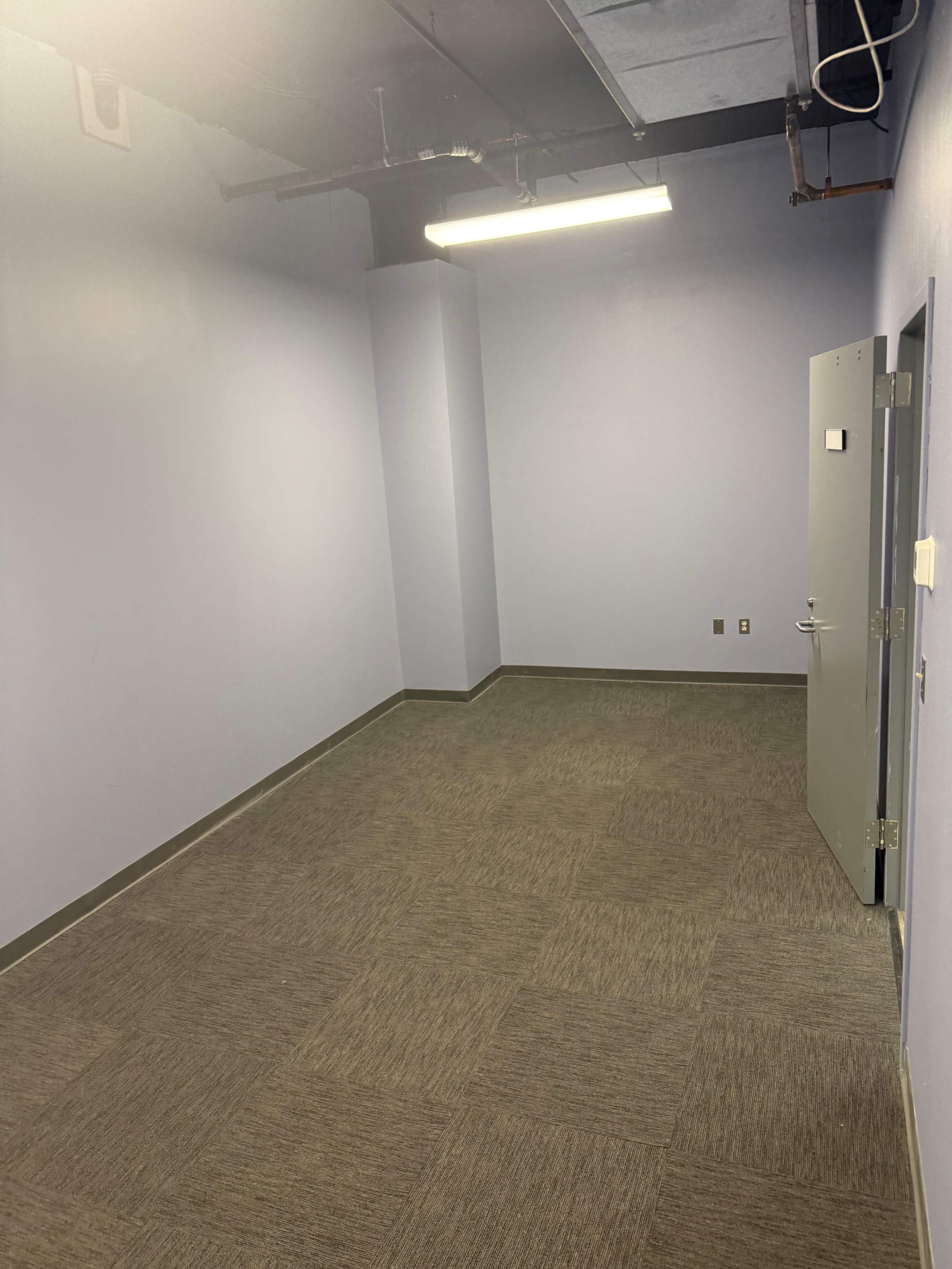 The image shows a bare, empty room with light purple walls and gray carpeting, featuring a single door on the right.