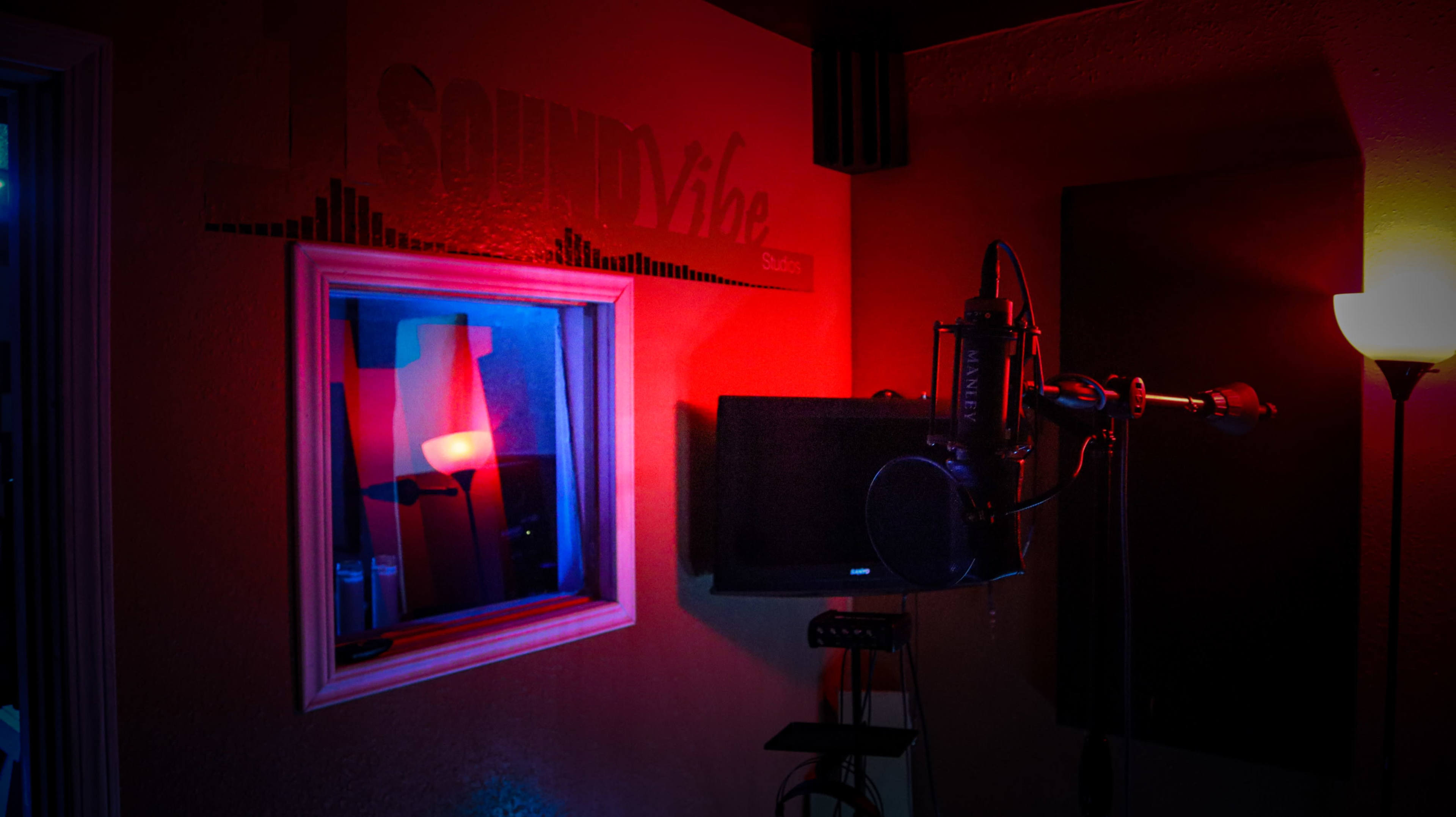A recording studio with a microphone, a monitor, and a window reflecting a red lamp.