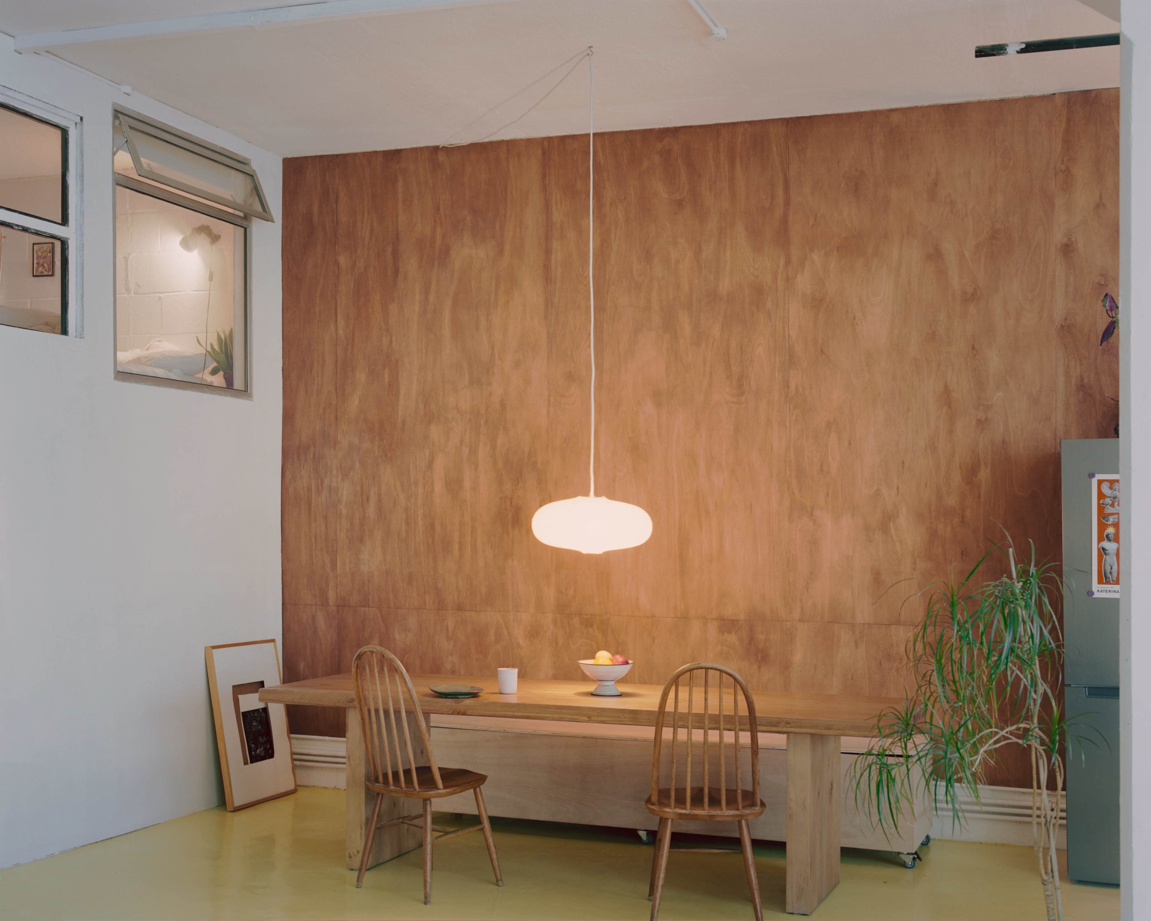 The image shows a minimalist dining area featuring a wooden table with two chairs, a pendant light hanging above, and a wall clad in wood paneling.