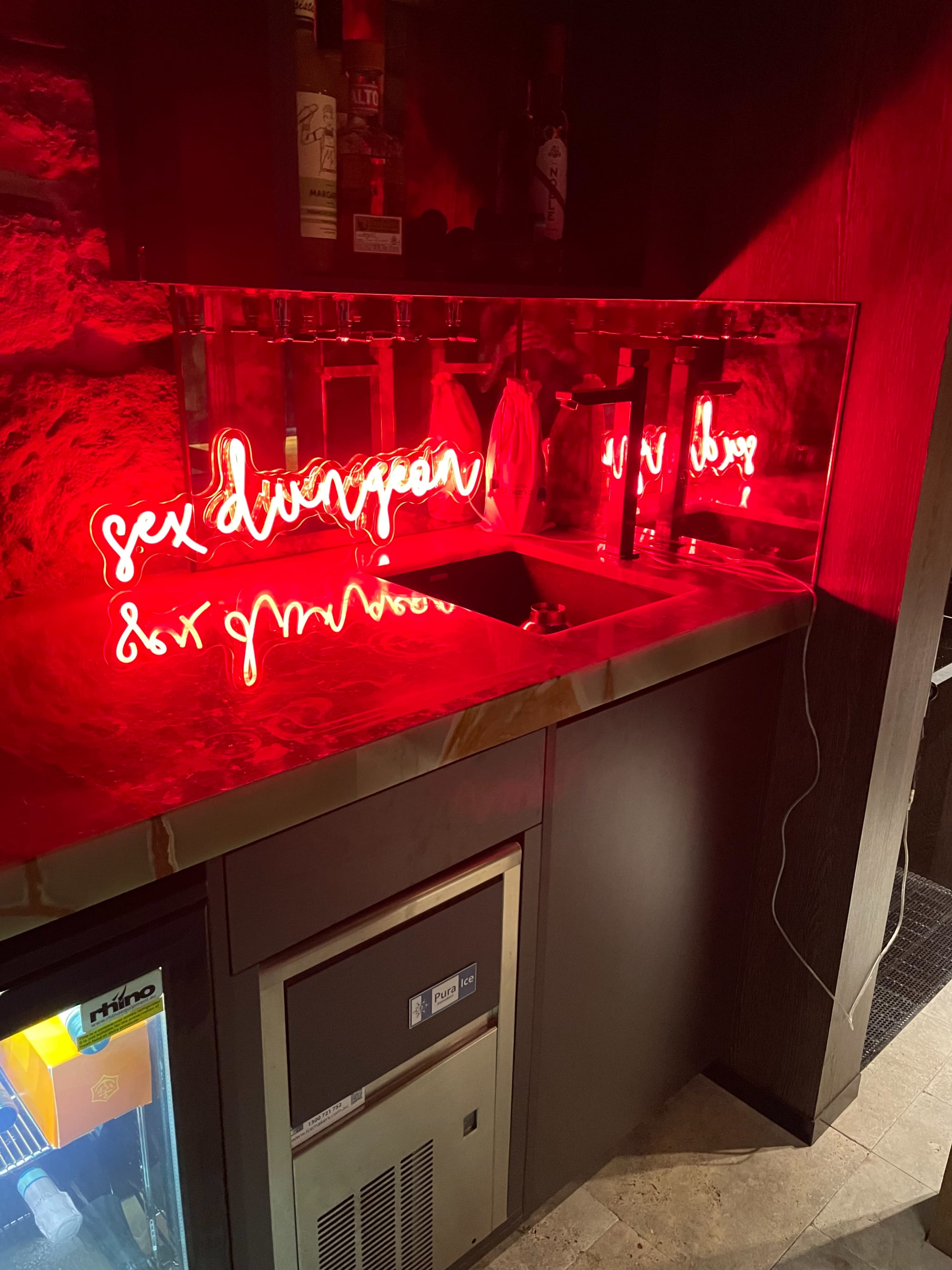 A bar counter features a glowing red neon sign with the words "sex dungeon" reflected in a mirrored surface.