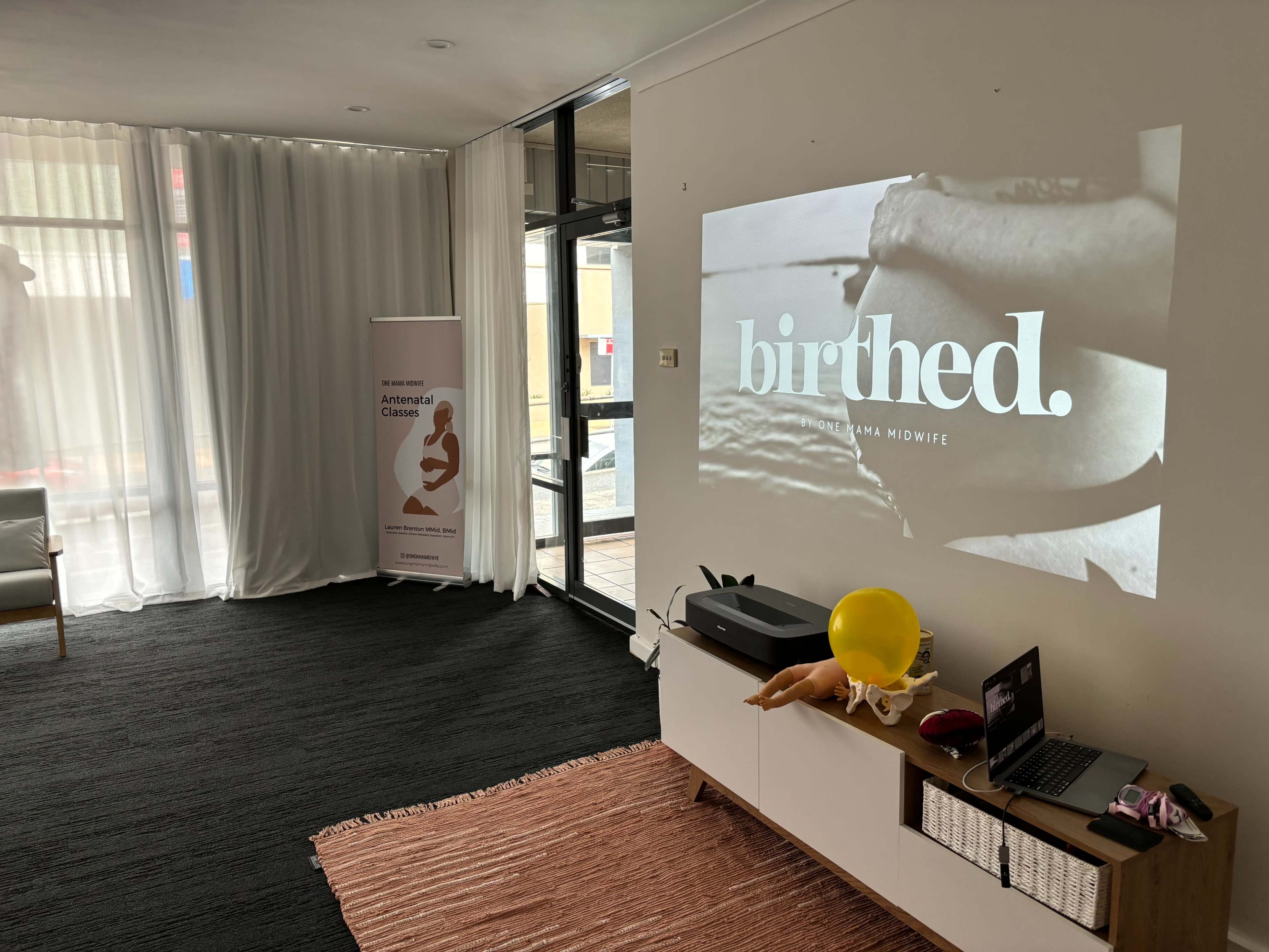 A projector displays the word "Birthed." on a wall in a room with light curtains, a banner, and various items on a side table.