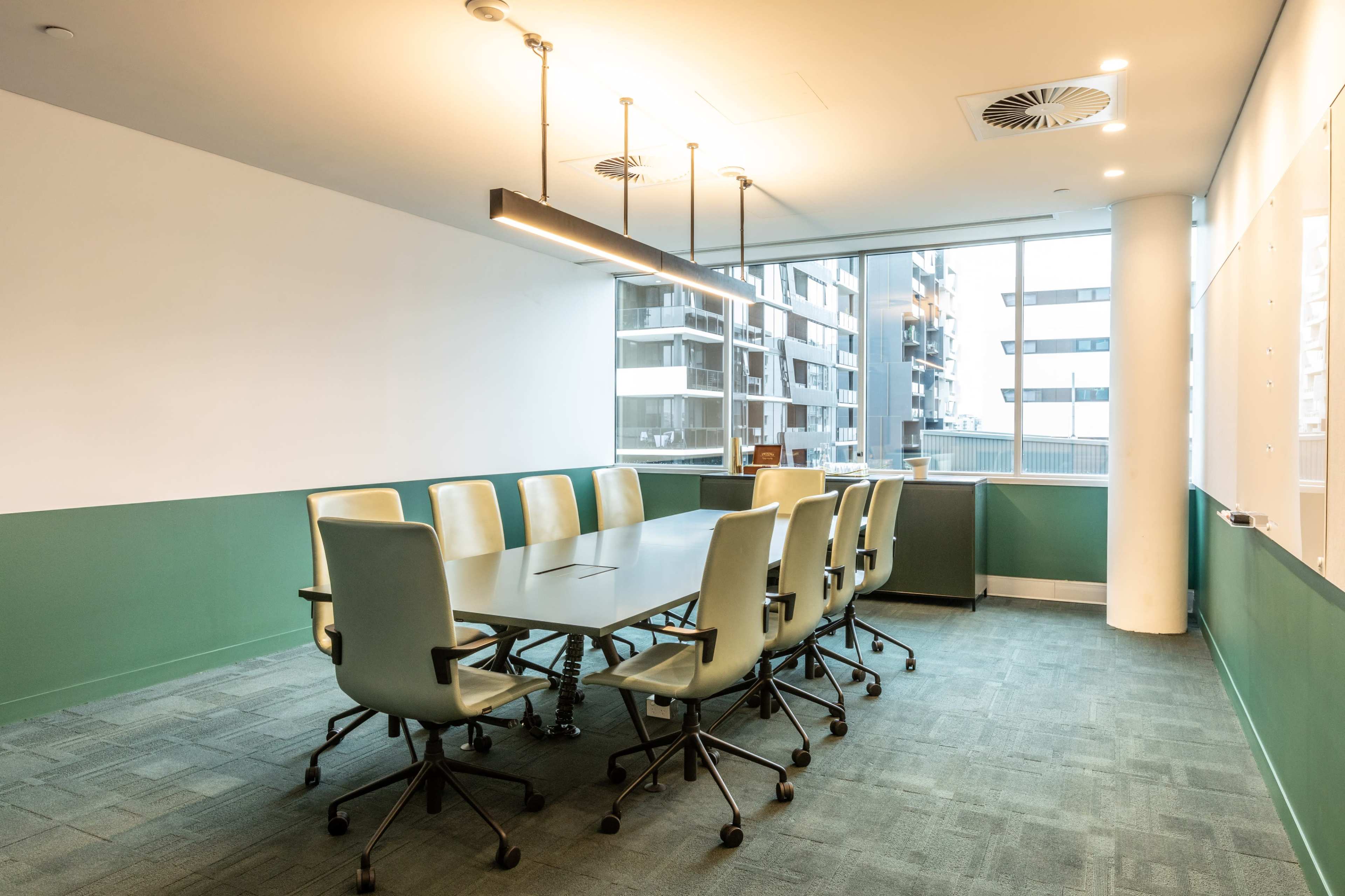 A modern conference room features a long table surrounded by ten green and white chairs, with large windows providing natural light.