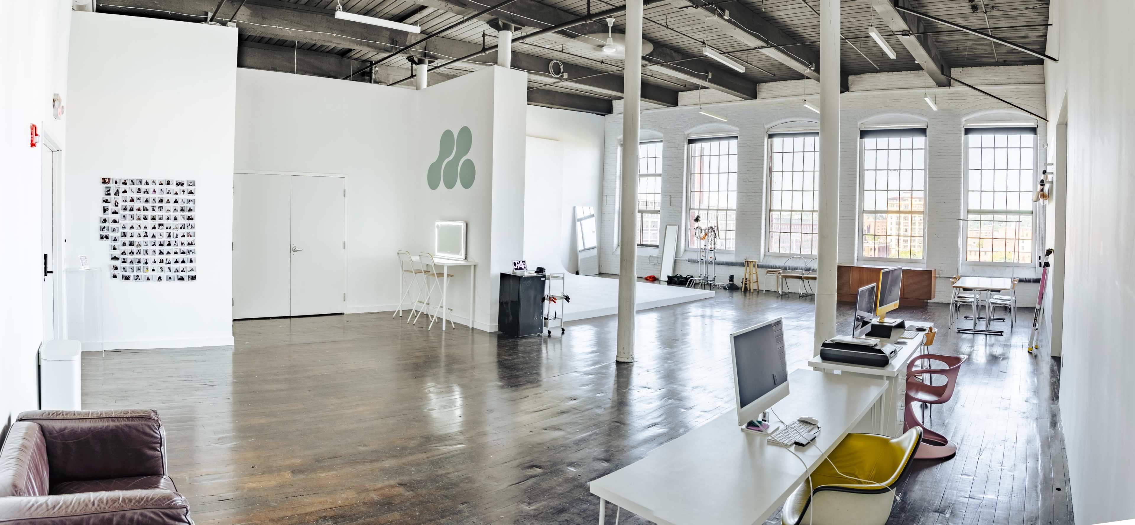 A spacious, well-lit studio with large windows, wooden floors, and a minimalist design featuring seating areas and workstations.
