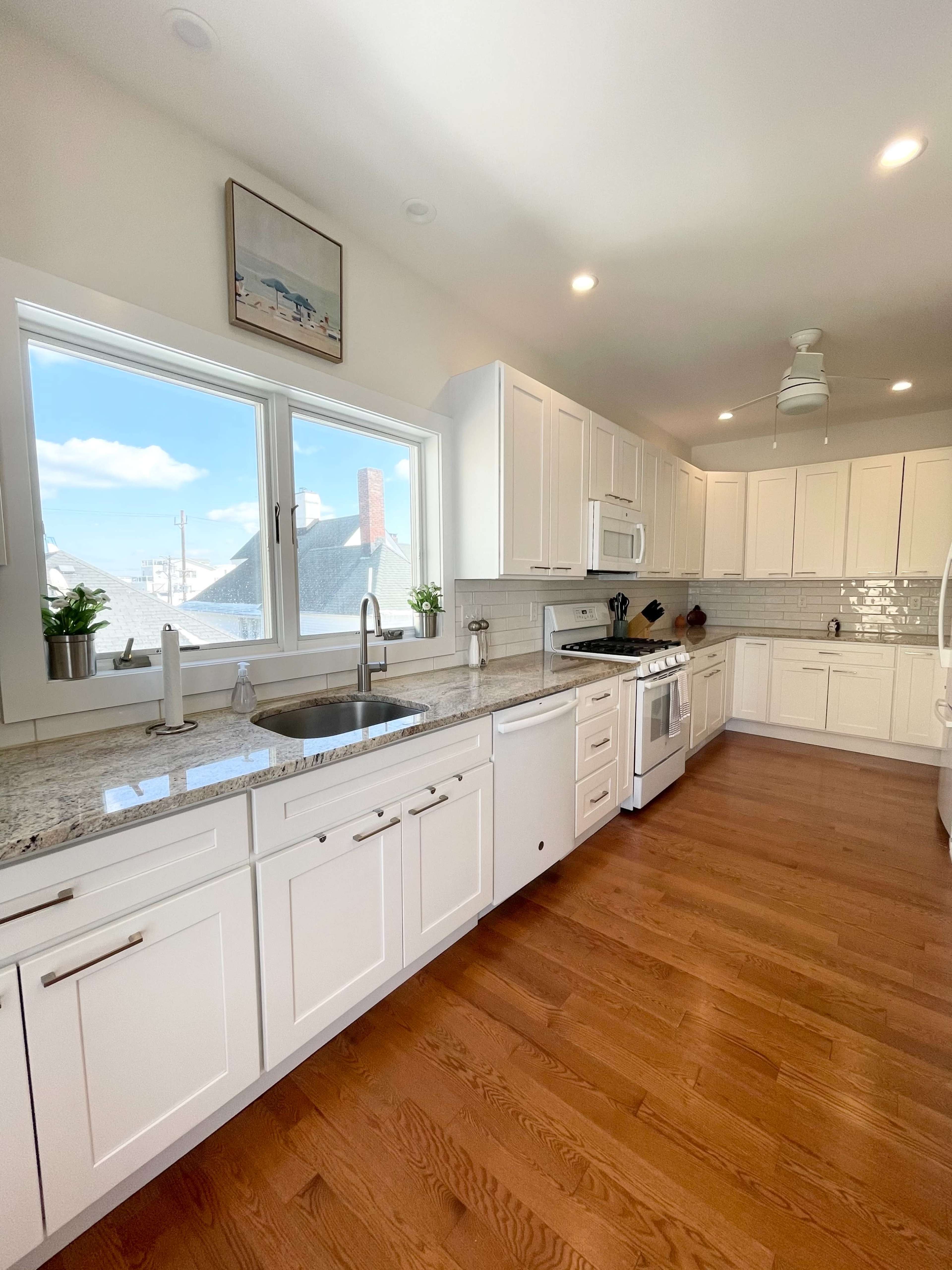 A modern kitchen features white cabinetry, a large window, hardwood floors, and stainless steel appliances.