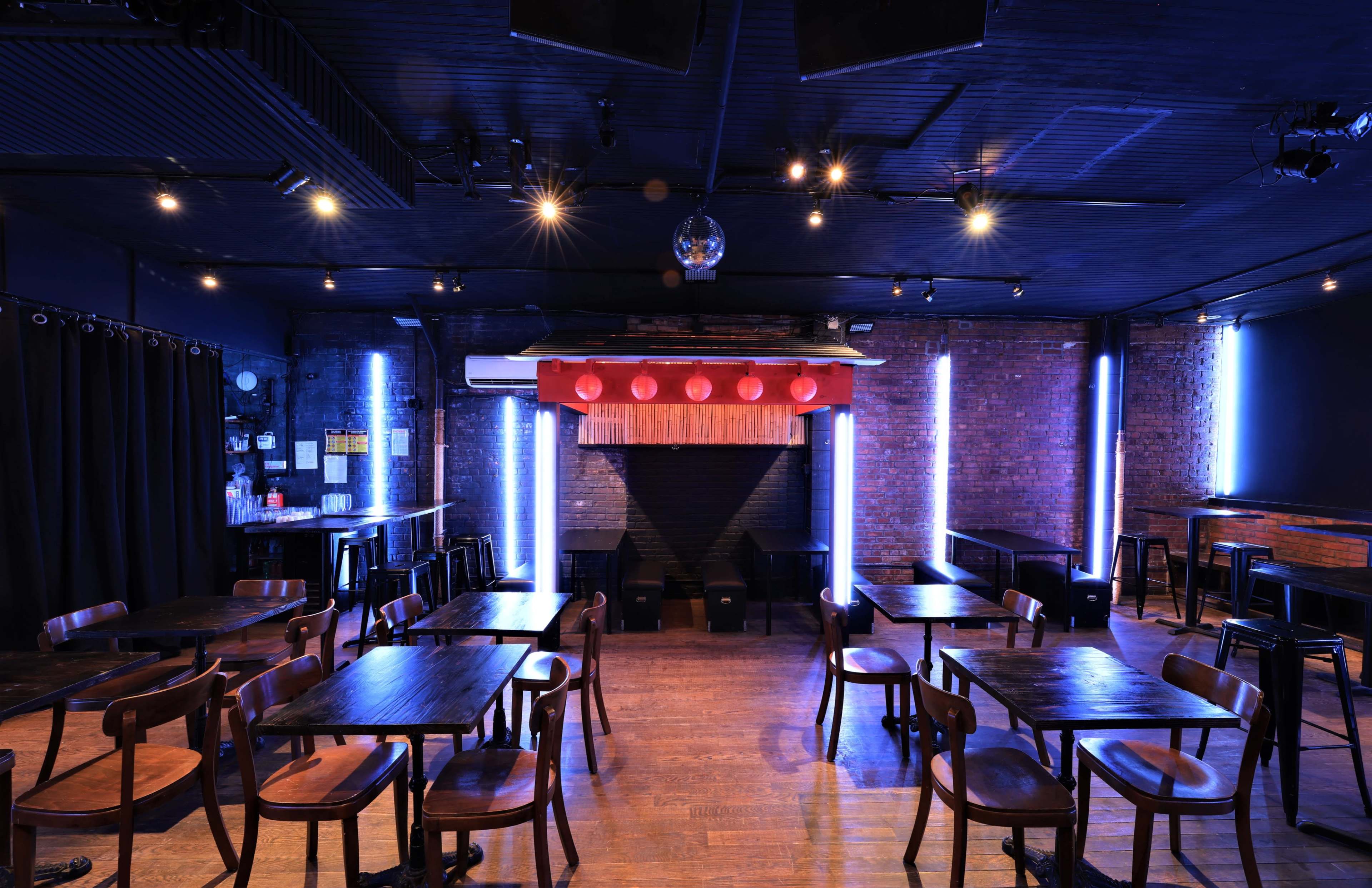 An empty bar or performance space with wooden tables and chairs, a brick wall, and red lanterns hanging above a stage area.