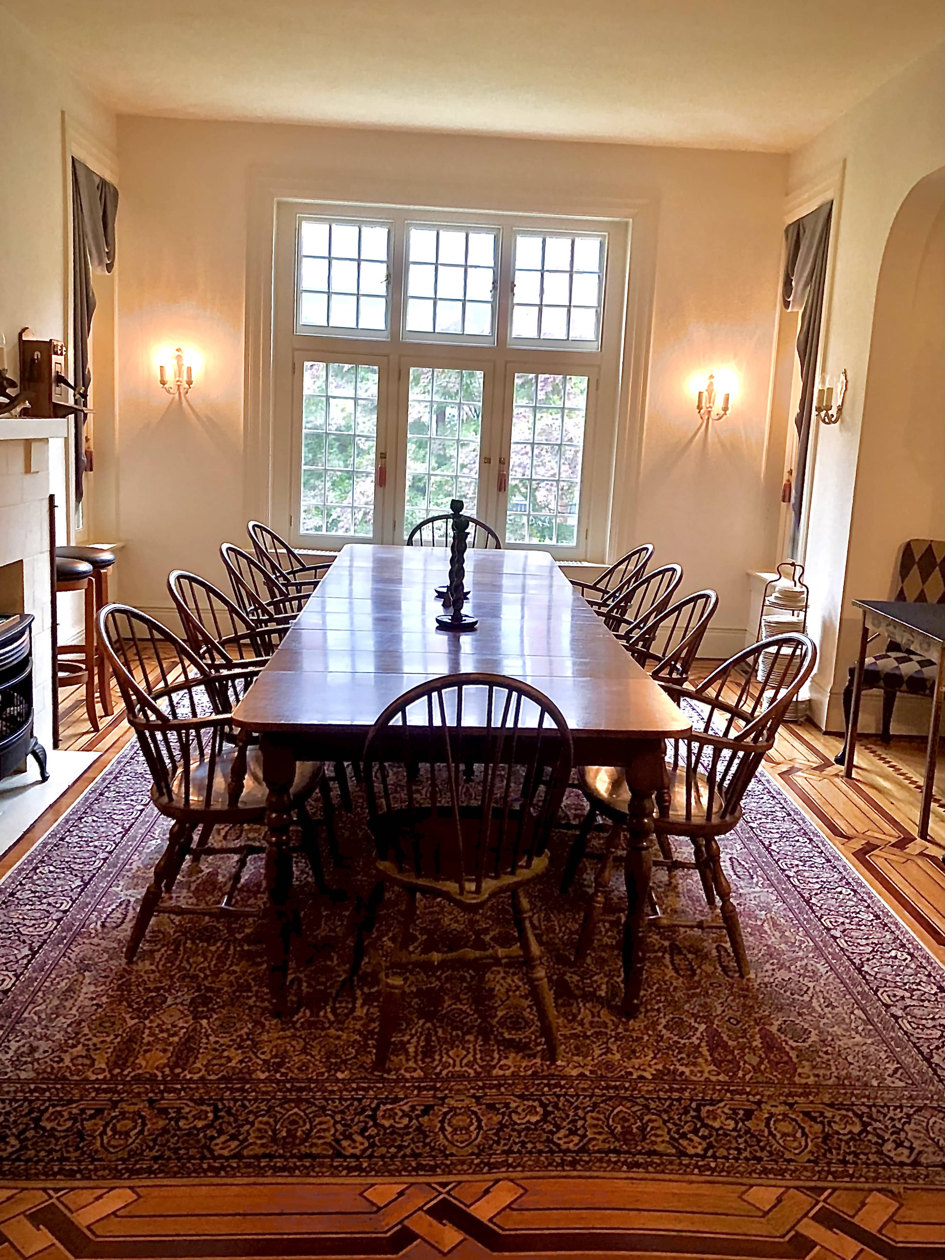 A large wooden dining table surrounded by ten chairs is positioned in a well-lit room with multiple windows and a decorative rug on the floor.