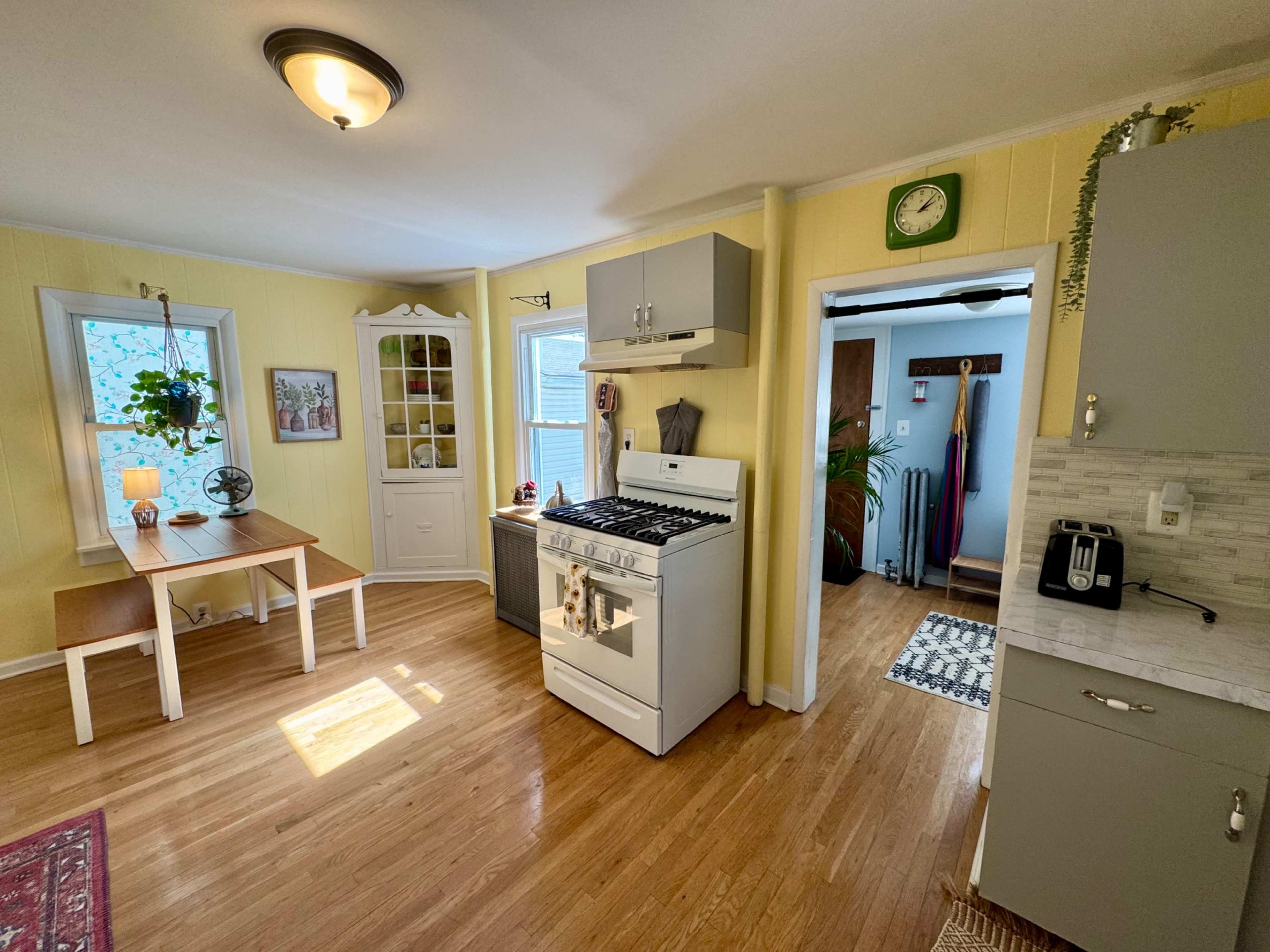 The image shows a kitchen with a gas stove, a small dining table, and cabinets against yellow walls, featuring bright natural light from a nearby window.