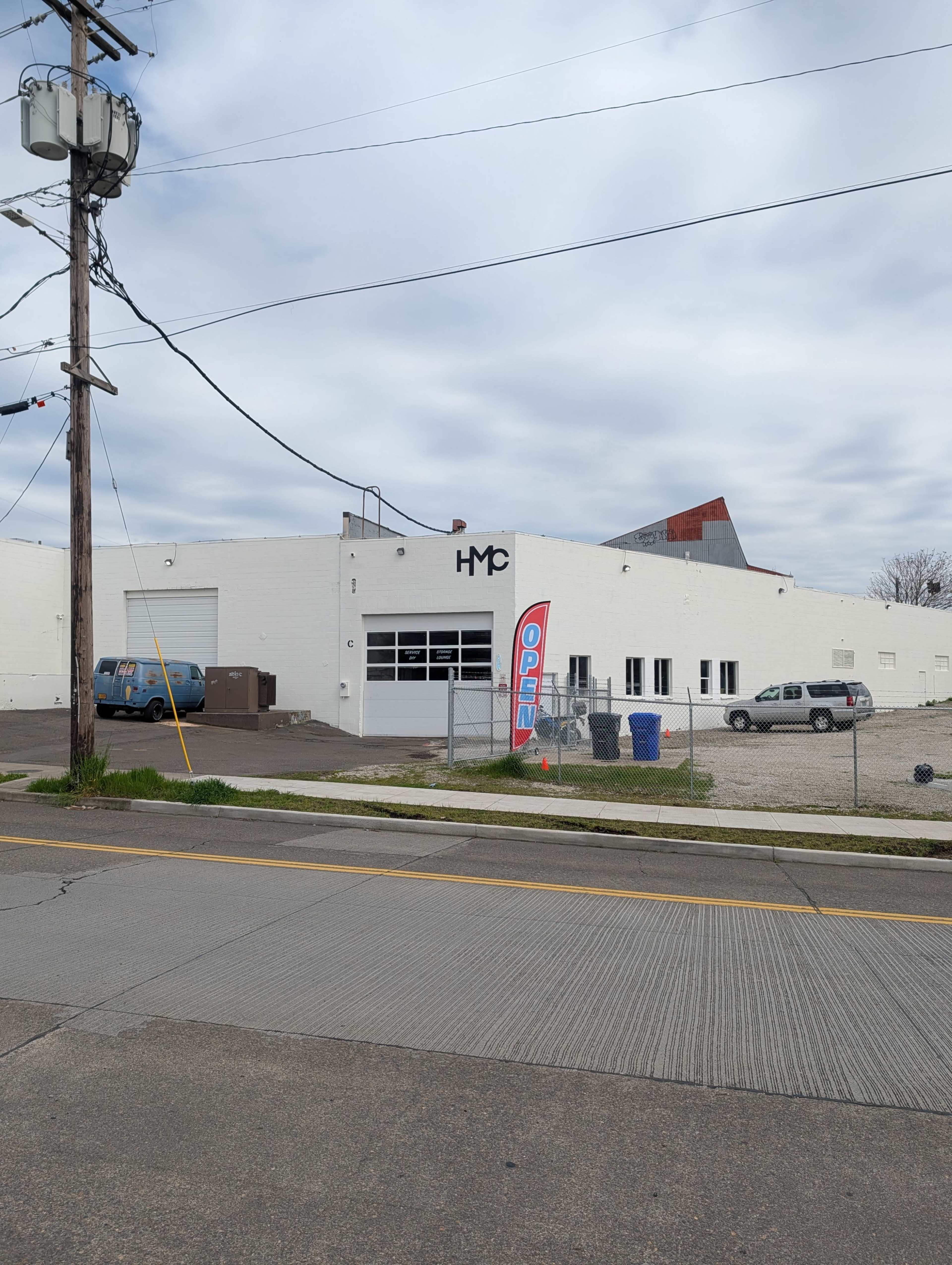 A white warehouse features large glass doors, a "OPEN" sign, and is situated next to a power pole and parked vehicles.
