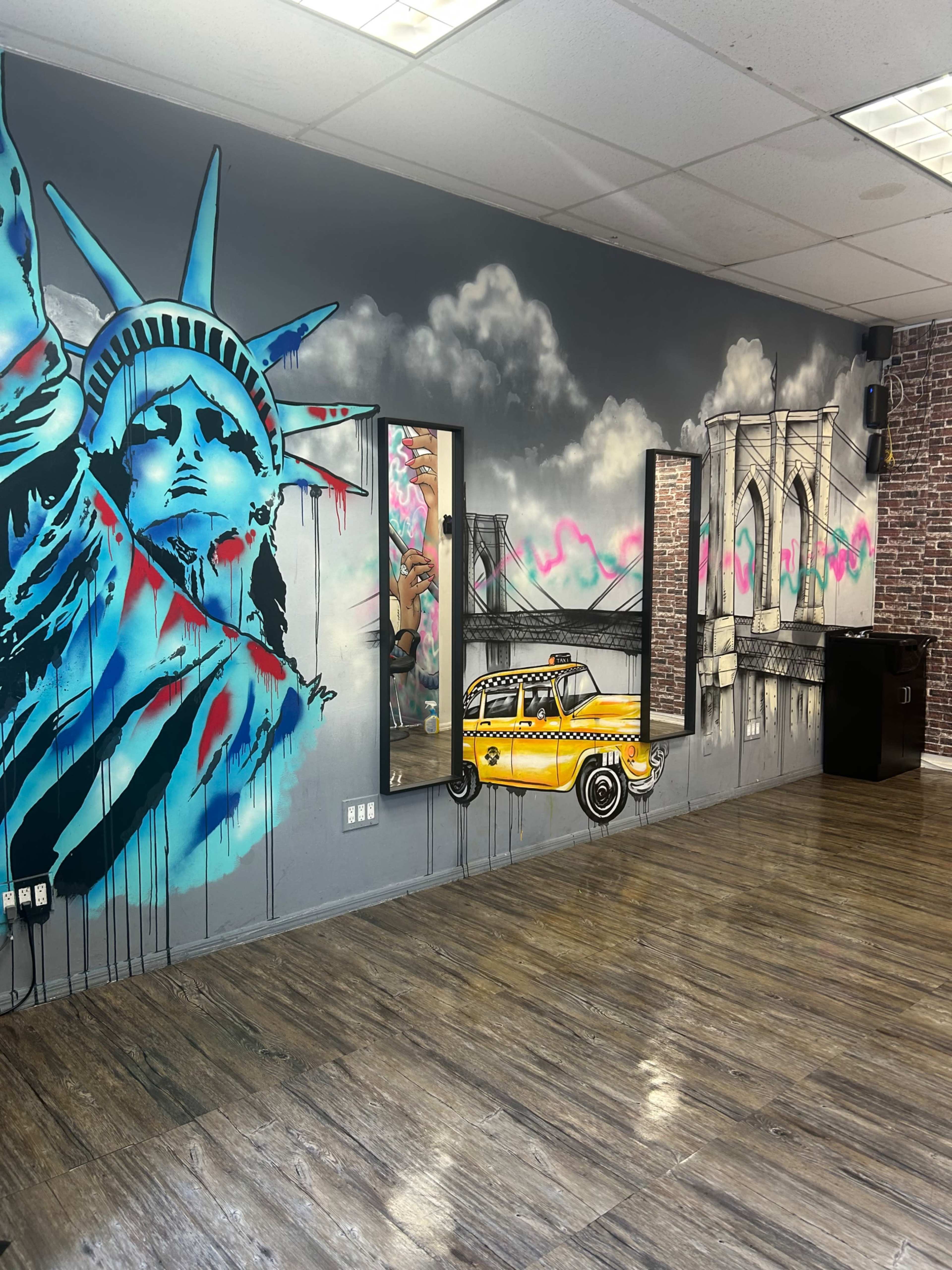 The image shows an interior space featuring a mural of the Statue of Liberty, a yellow taxi, and the Brooklyn Bridge on a grey wall, with wooden flooring and mirrors visible.