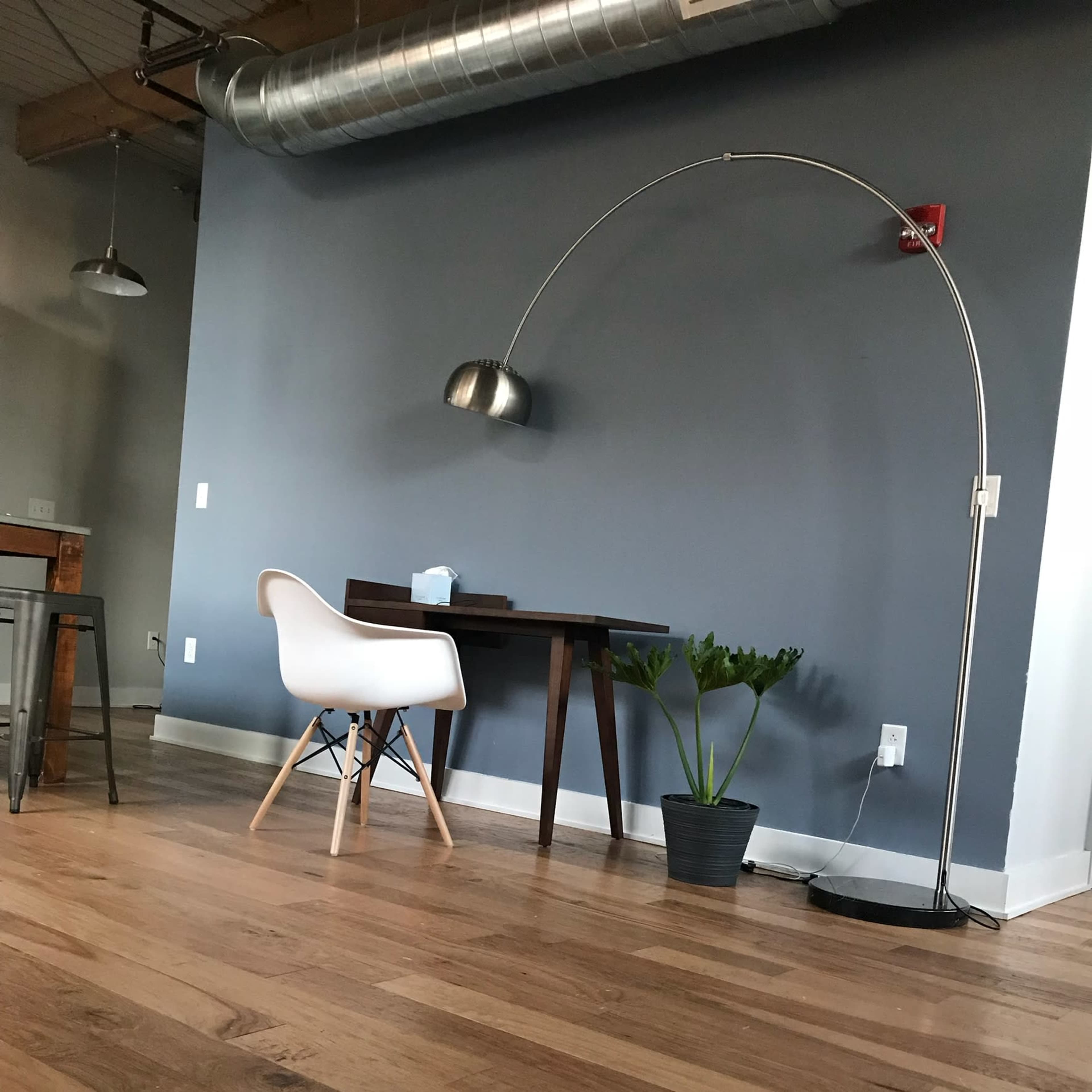 A modern workspace features a sleek desk, an arc floor lamp, and a potted plant against a gray wall with wooden flooring.