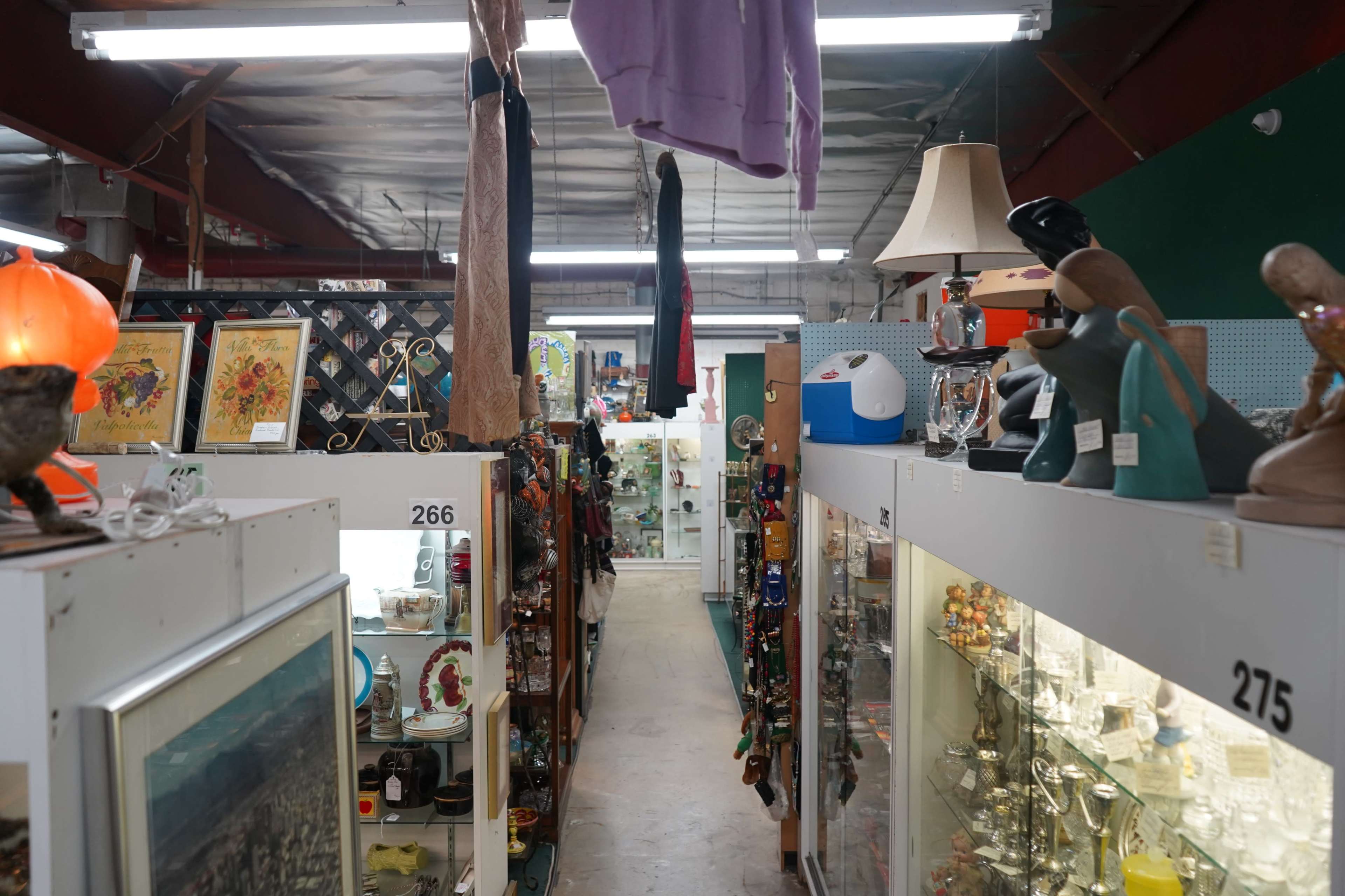 The image shows the interior of an antique store with narrow aisles lined with various shelves displaying diverse items such as artwork, clothing, and home decor.
