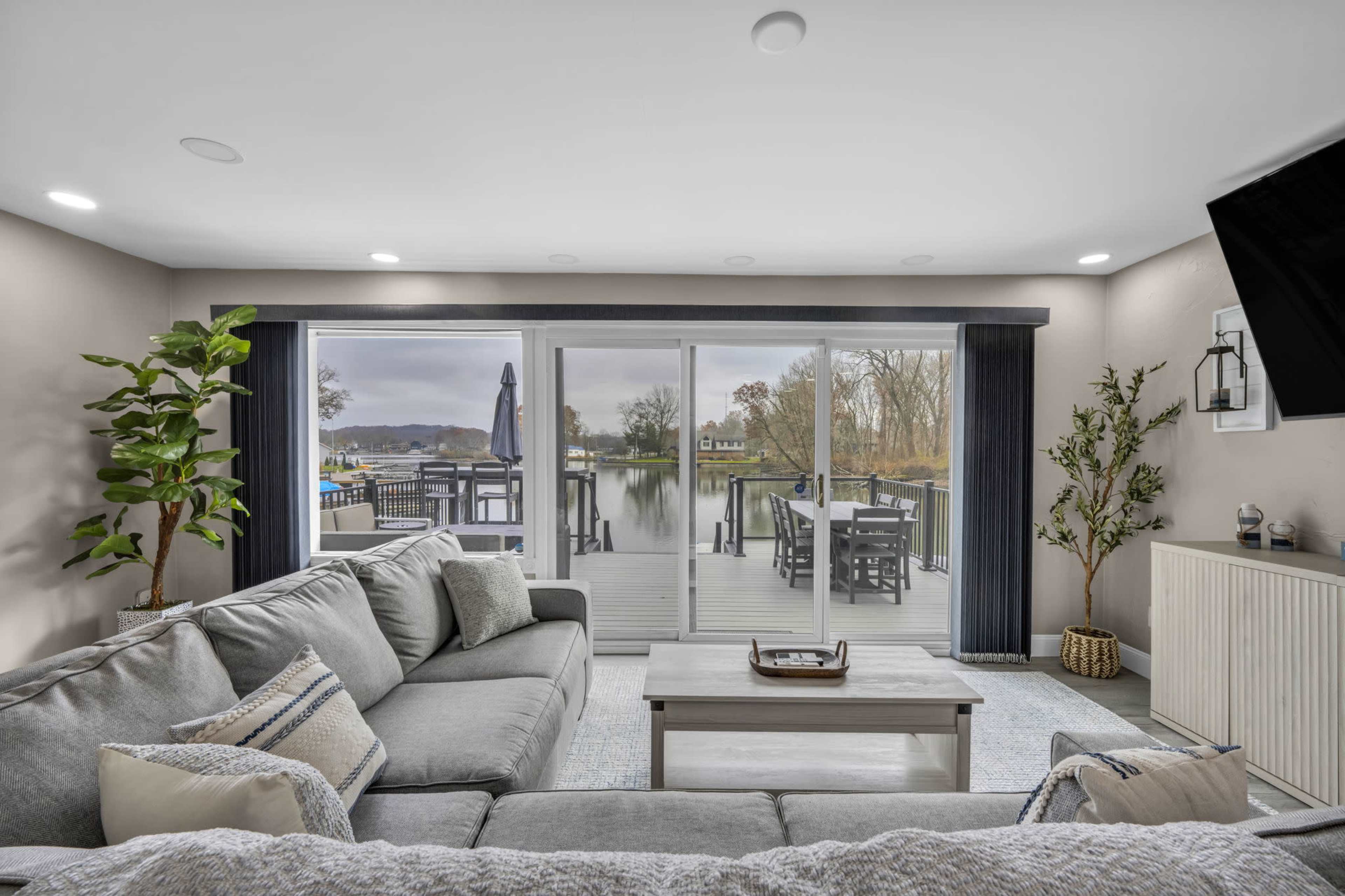 The image shows a modern living room with a gray sofa, a coffee table, and large sliding glass doors opening to a deck overlooking a body of water.