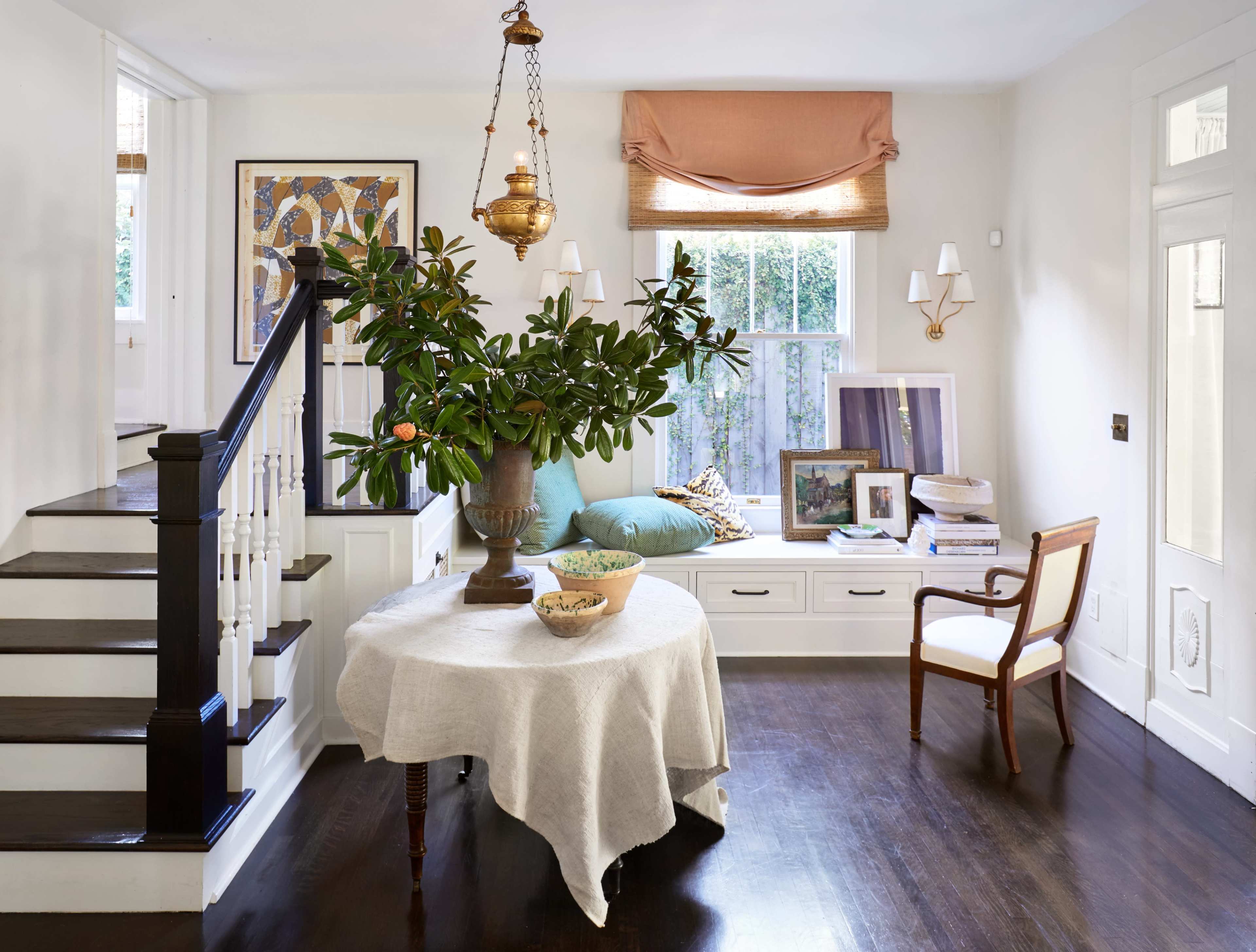 The image shows a bright, airy living space featuring a round table with a tablecloth, a staircase, a window seat with decorative pillows, and a potted plant on the table.