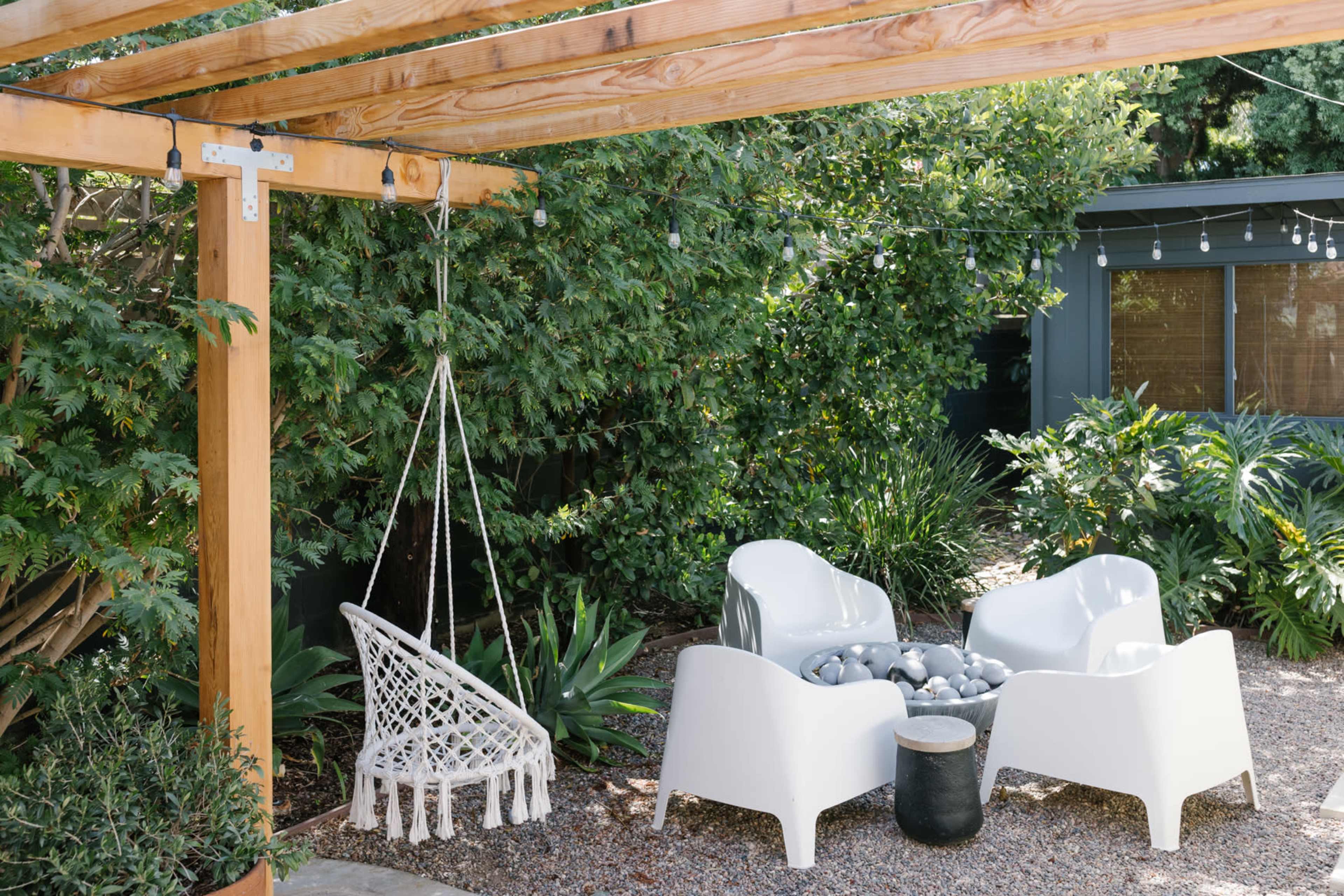 A cozy outdoor seating area features a hanging chair and four white chairs arranged around a small table, surrounded by lush greenery.