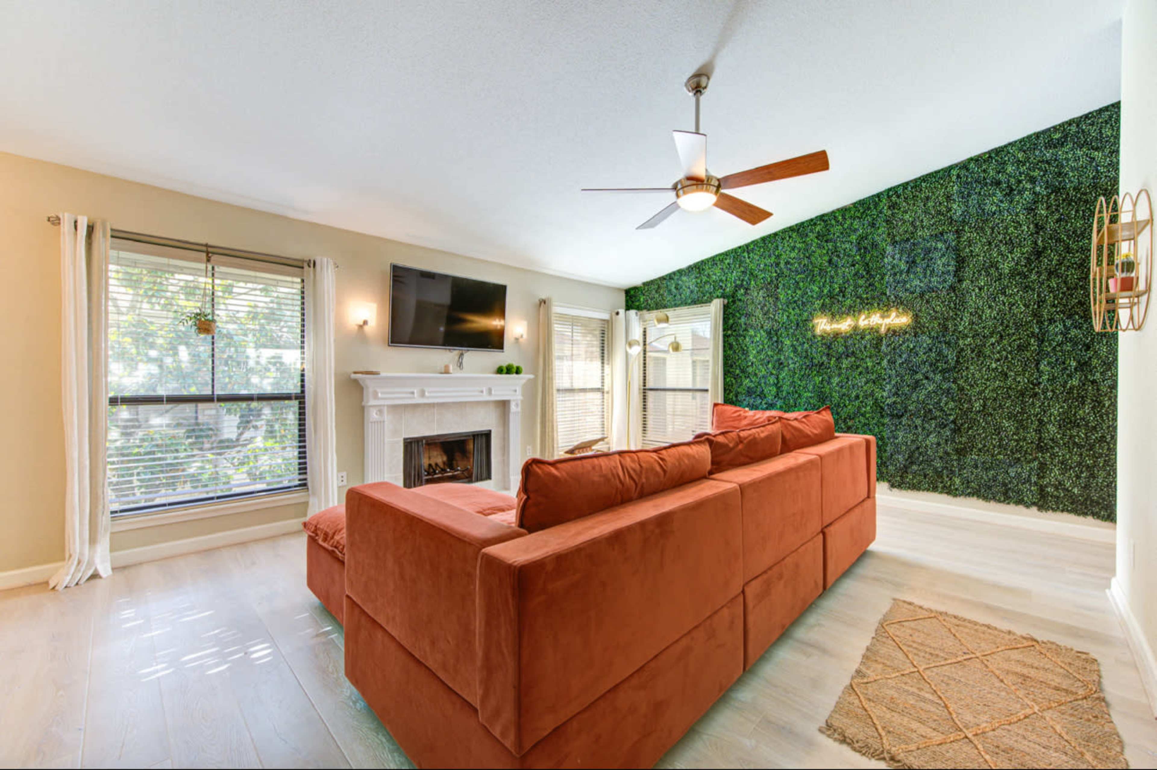 A modern living room features a large couch, a flat-screen TV mounted on a wall, and a greenery accent wall beside a fireplace.