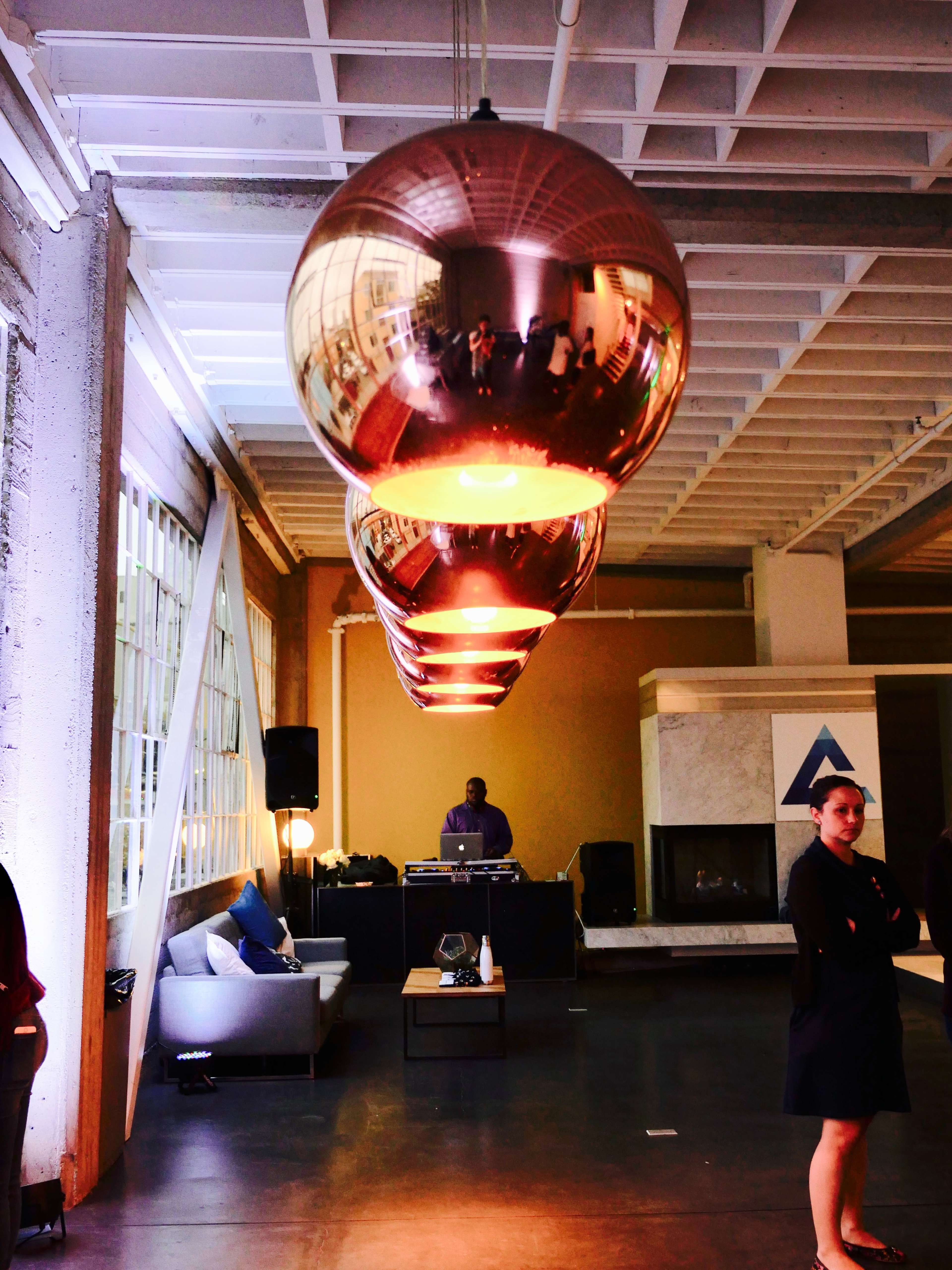 A modern indoor event space featuring four large, reflective spherical pendant lights hanging in a row, while a DJ is set up in the background and a woman stands to the side.