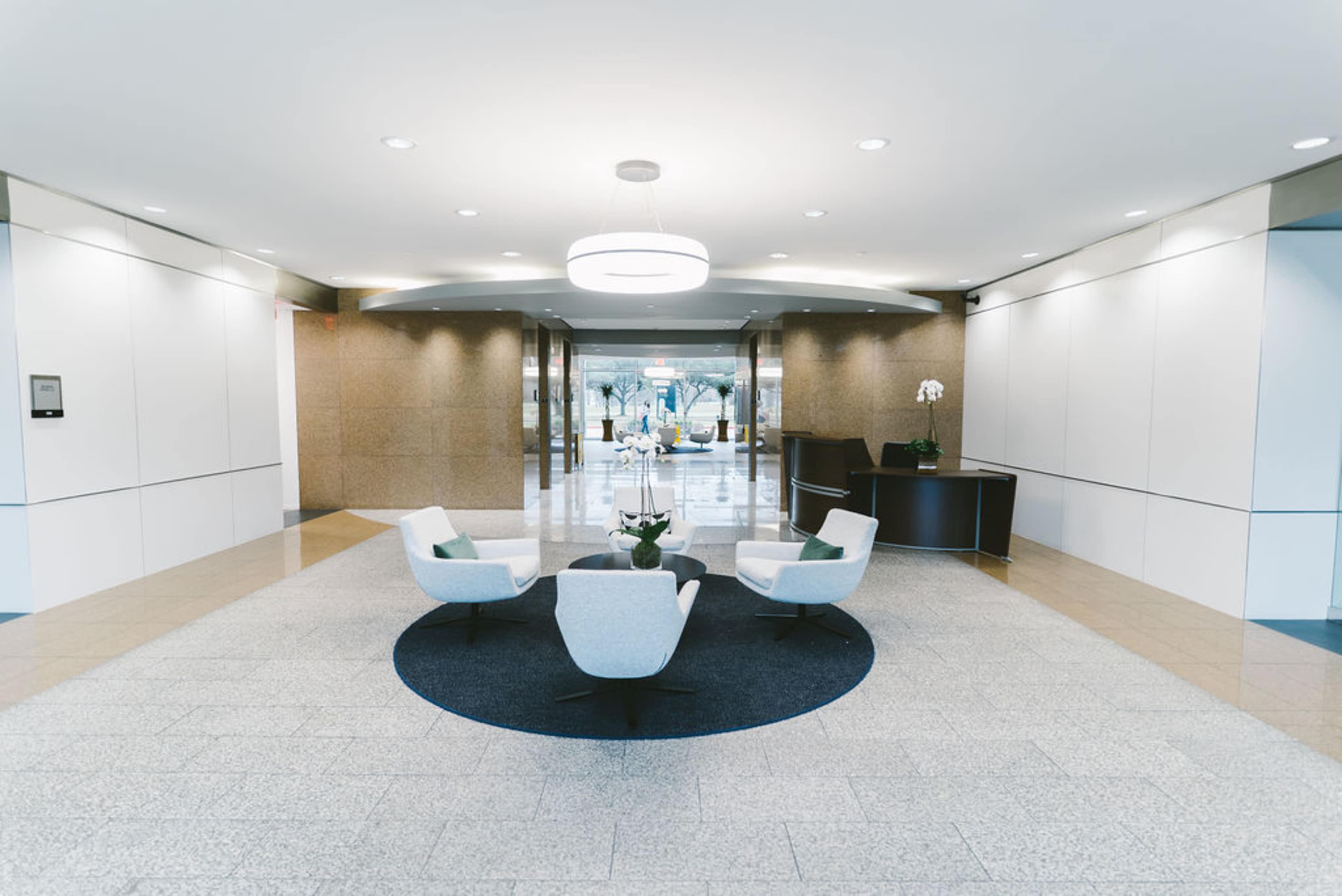 The image shows a modern lobby featuring a circular seating arrangement with three chairs on a round rug, a reception desk to the right, and a large entrance ahead.