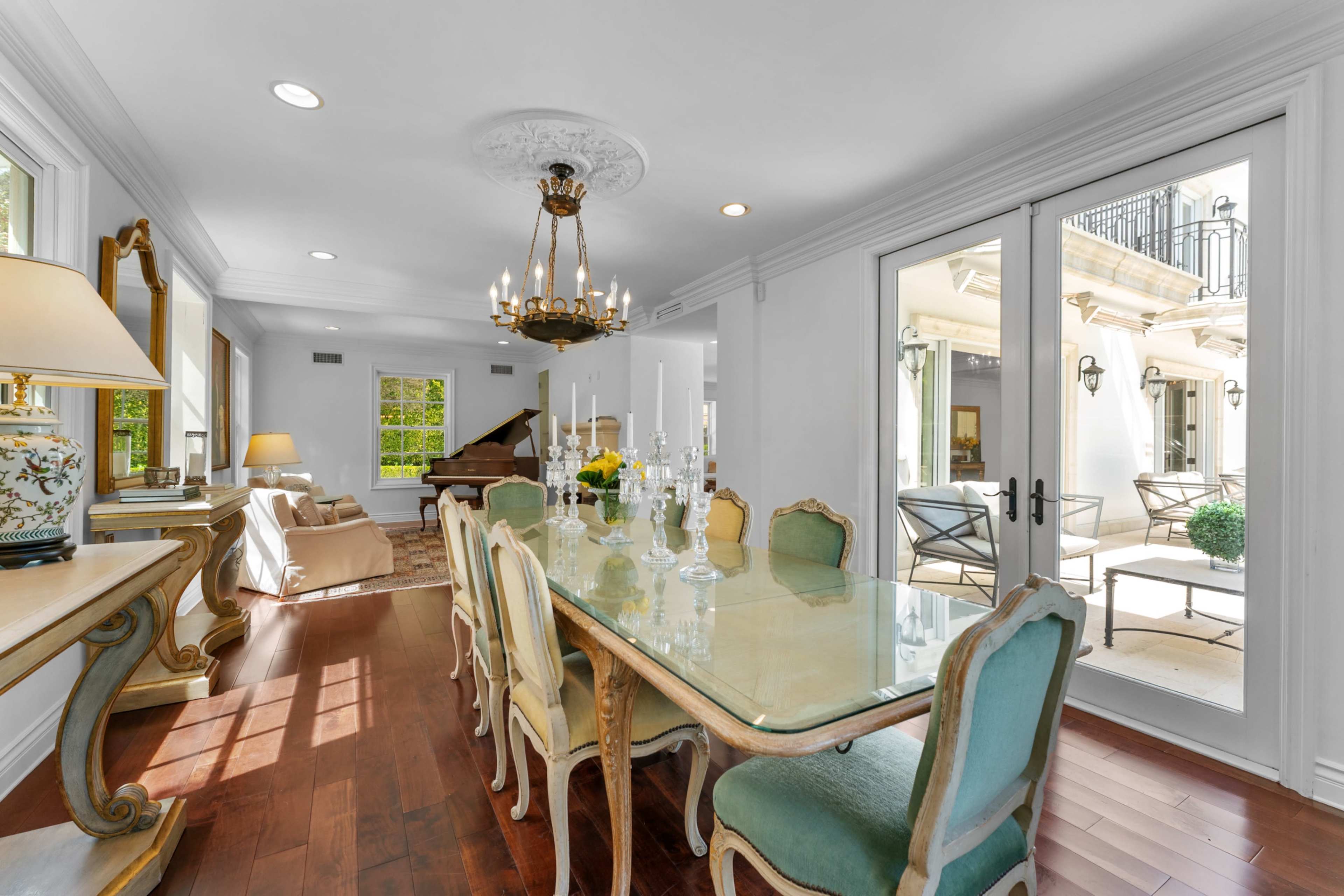 The image shows a bright dining area with a long glass table surrounded by light-colored chairs, a chandelier above, and large windows opening to an outdoor patio.
