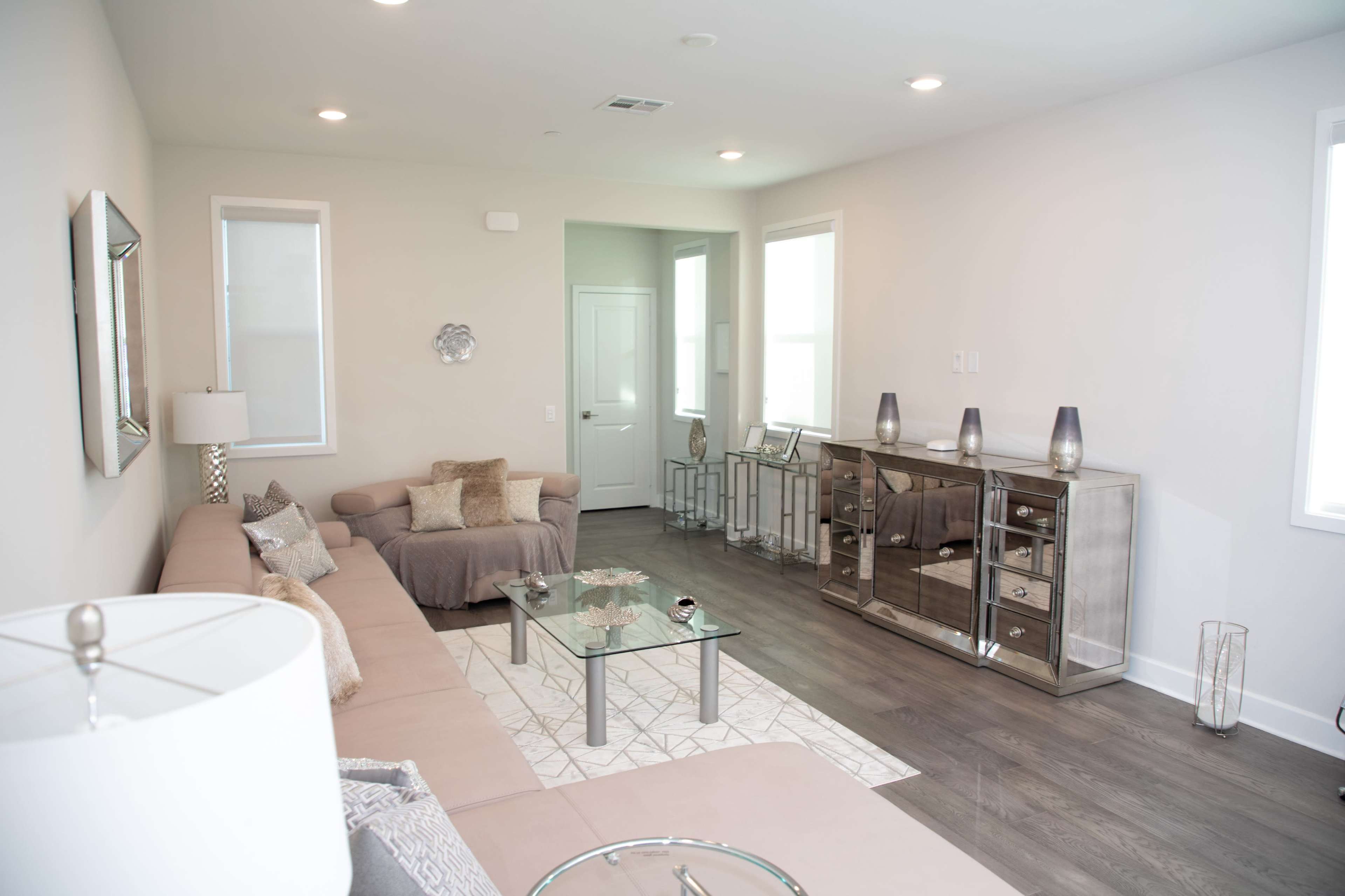 The room features a light-colored sofa, a glass coffee table, and a mirrored sideboard against the wall, with large windows allowing natural light to enter.