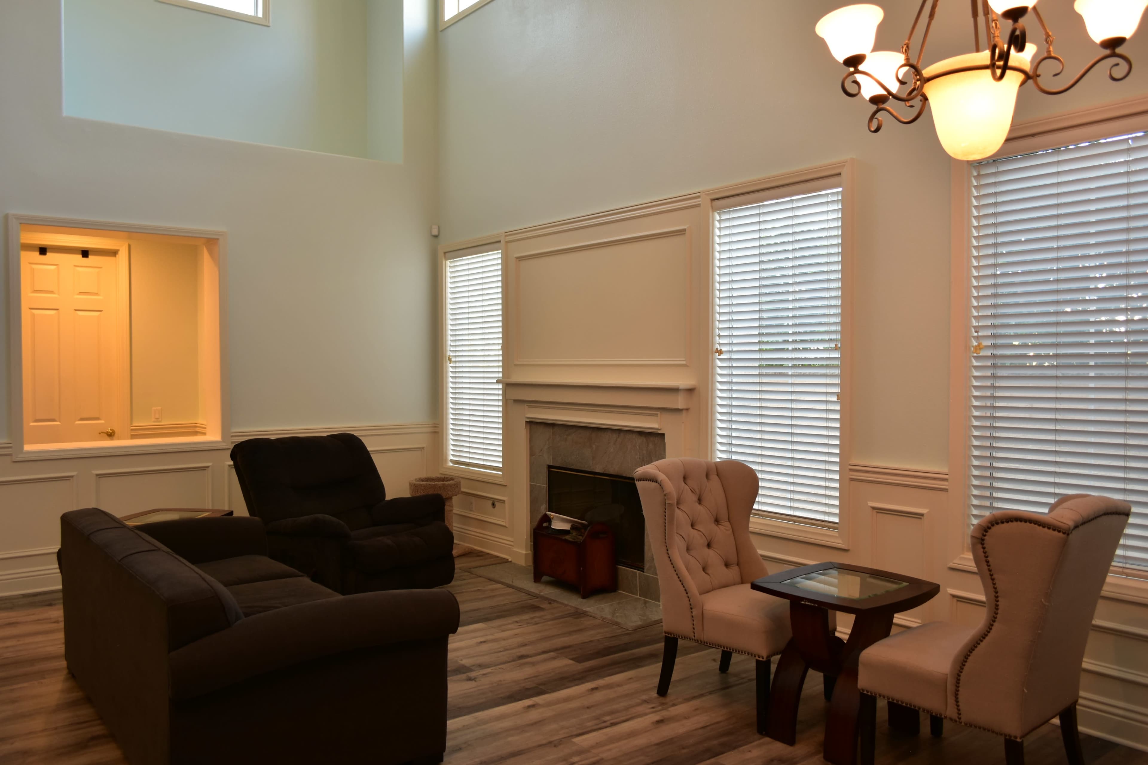 A spacious living room features a gray sofa, two armchairs, a small table, and a fireplace with large windows letting in natural light.
