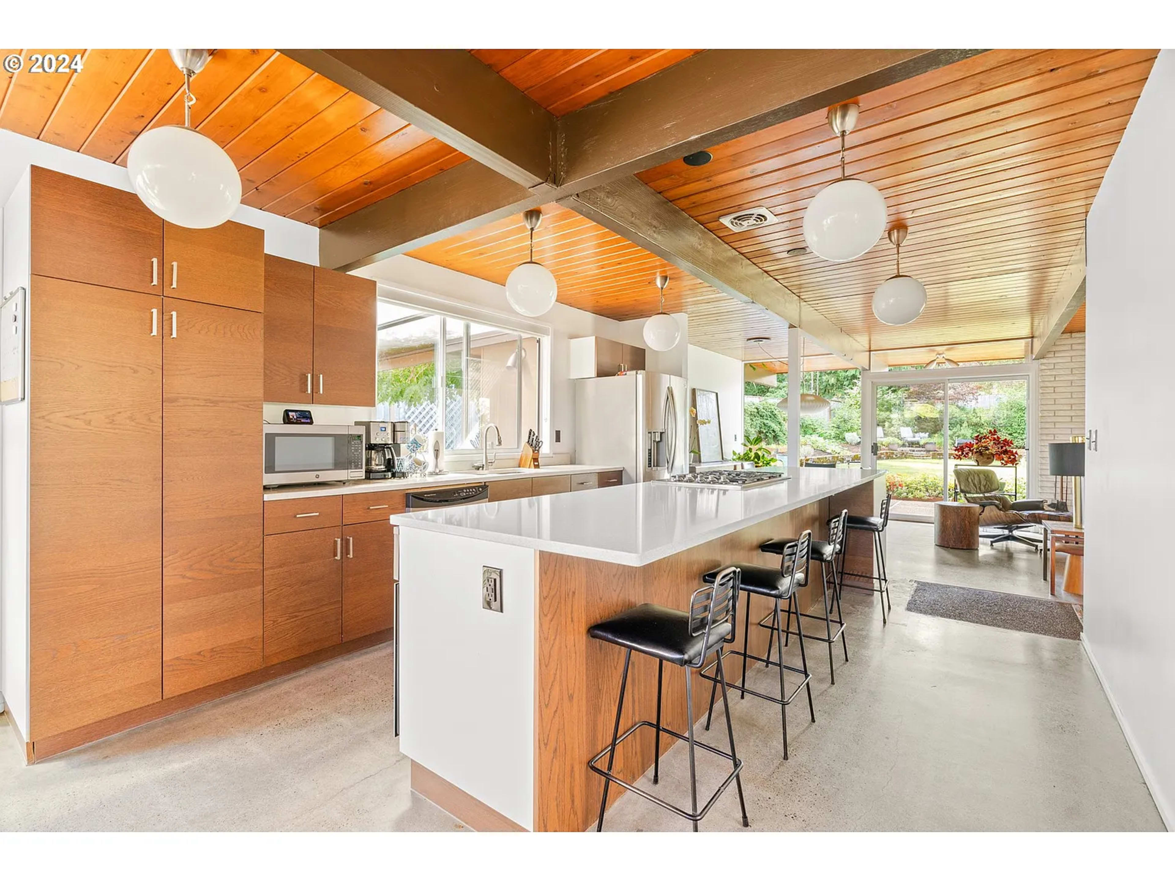 The image shows a modern kitchen featuring wooden cabinetry, a large island with bar seating, and large windows allowing natural light to fill the space.