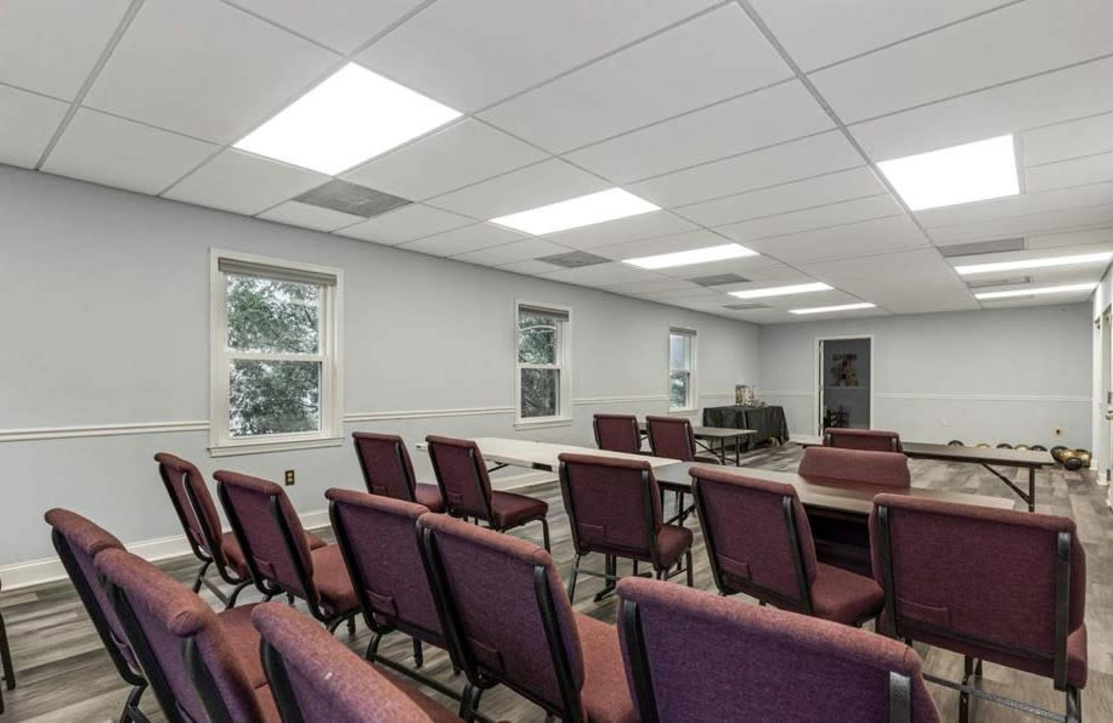 A meeting room with rows of maroon chairs arranged in front of tables, and several windows allowing natural light to enter.