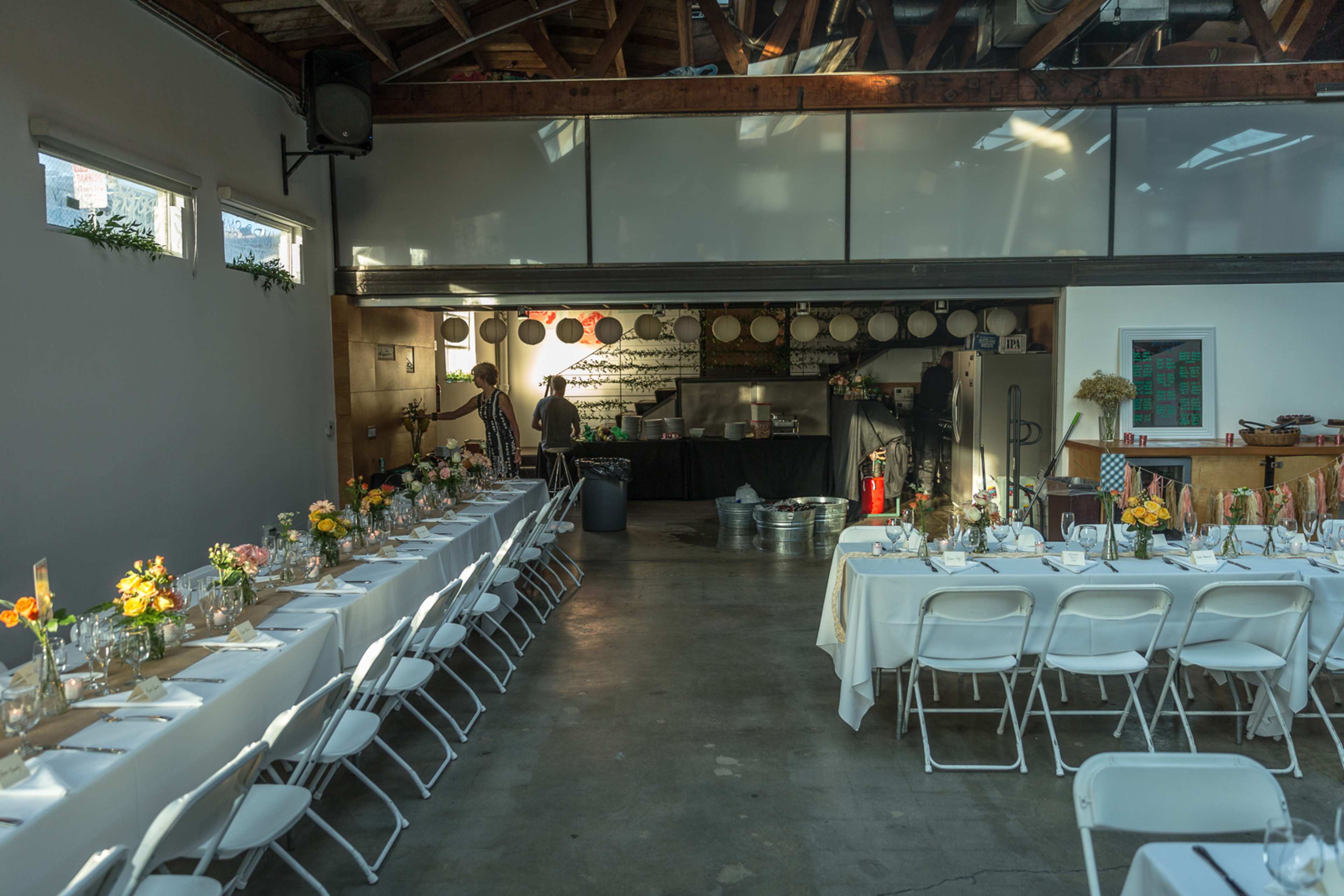 The image shows a spacious indoor venue set for an event, featuring long tables adorned with flowers and a kitchen area in the background.
