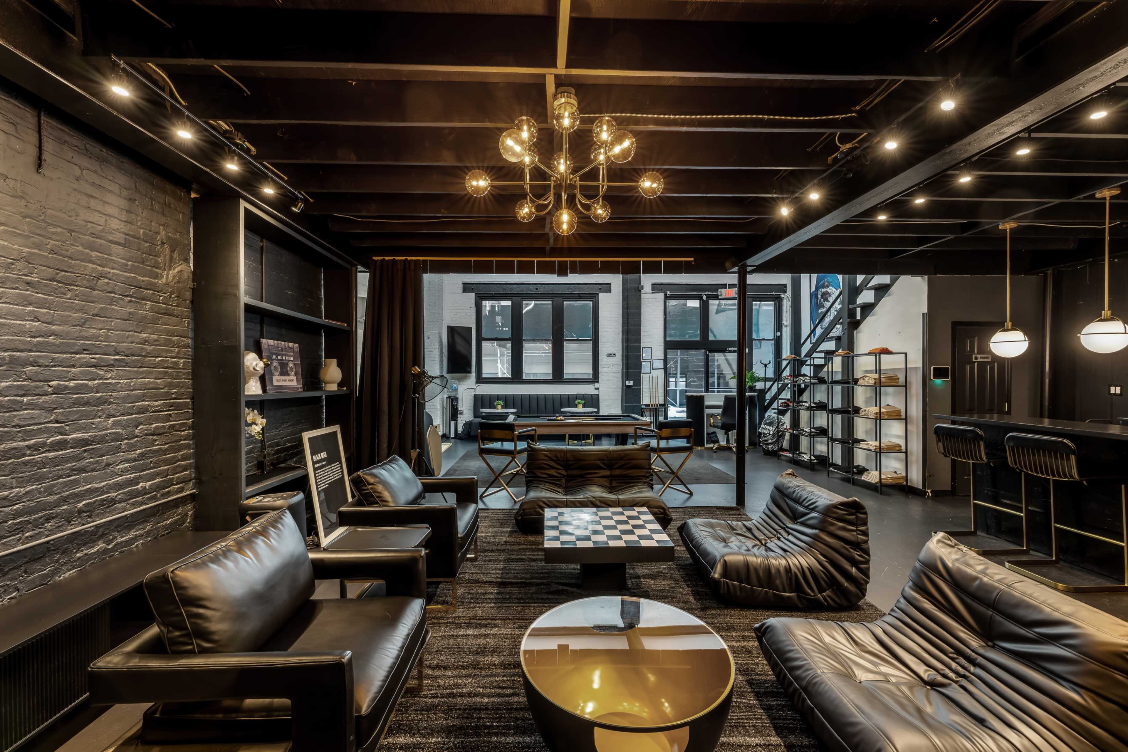 The image shows a modern interior space with black leather seating, a coffee table, large windows, and an open layout featuring a dining area and bookshelves.