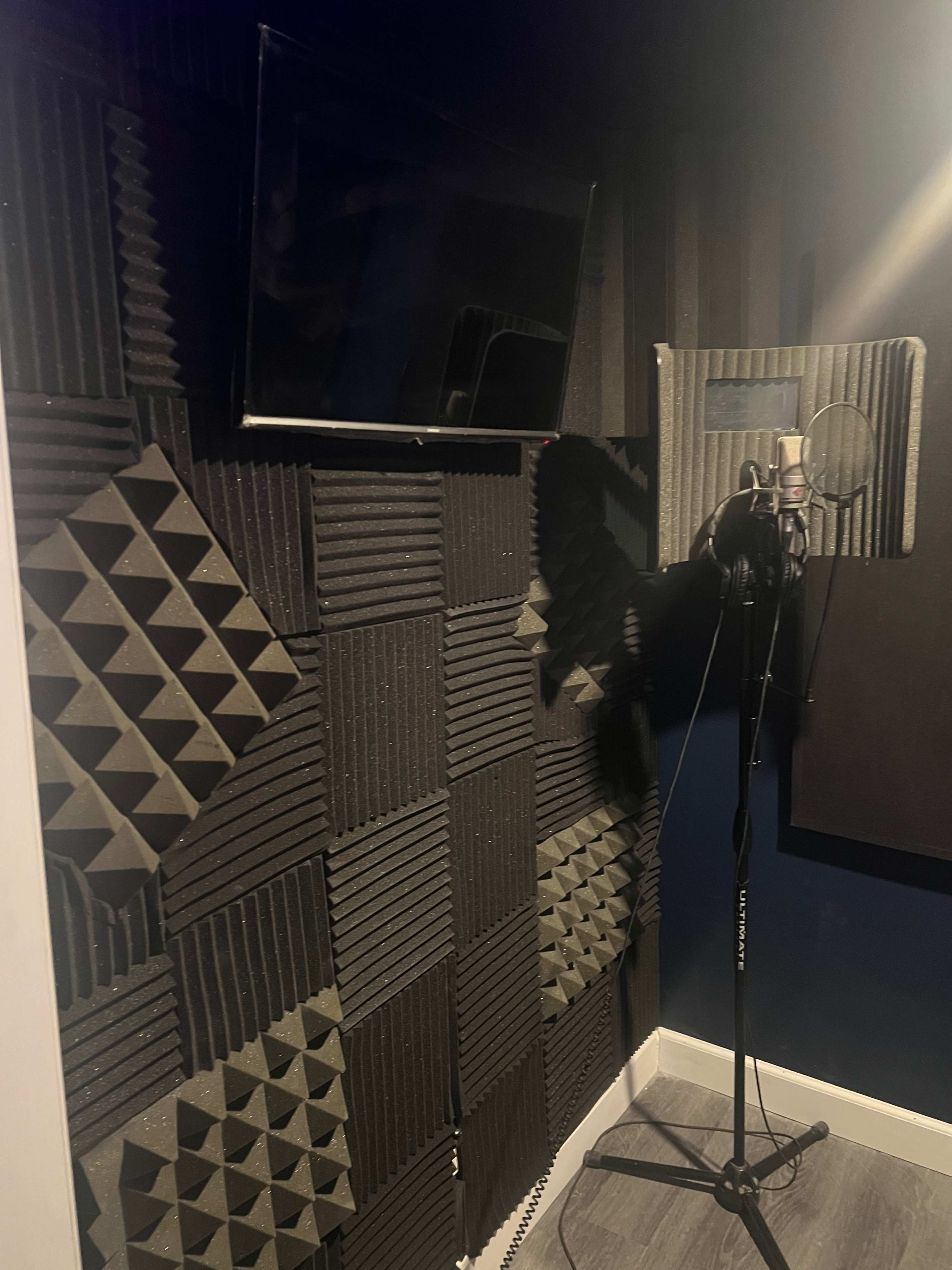 A soundproof recording studio featuring acoustic foam panels on the walls, a microphone on a stand, and a television mounted on the wall.