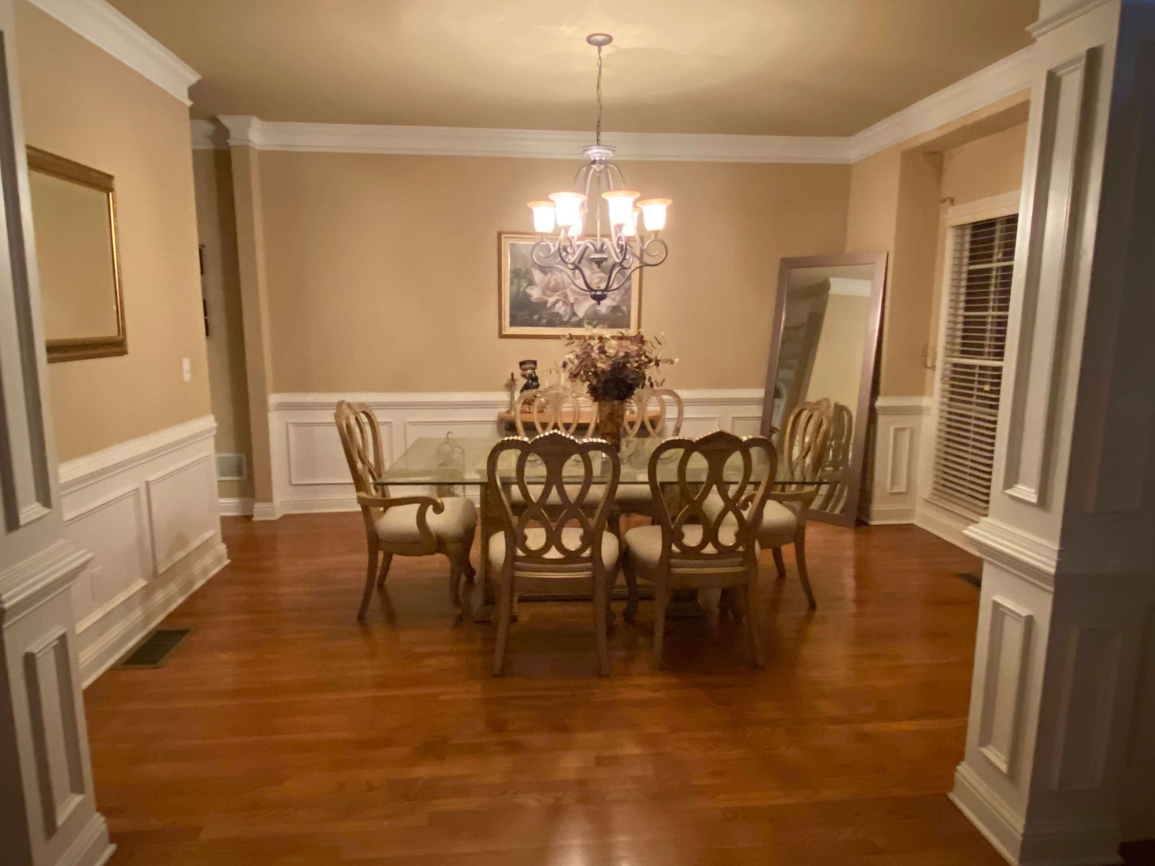 A dining room features a wooden table surrounded by chairs, illuminated by a chandelier, with a mirror and a framed painting on the walls.
