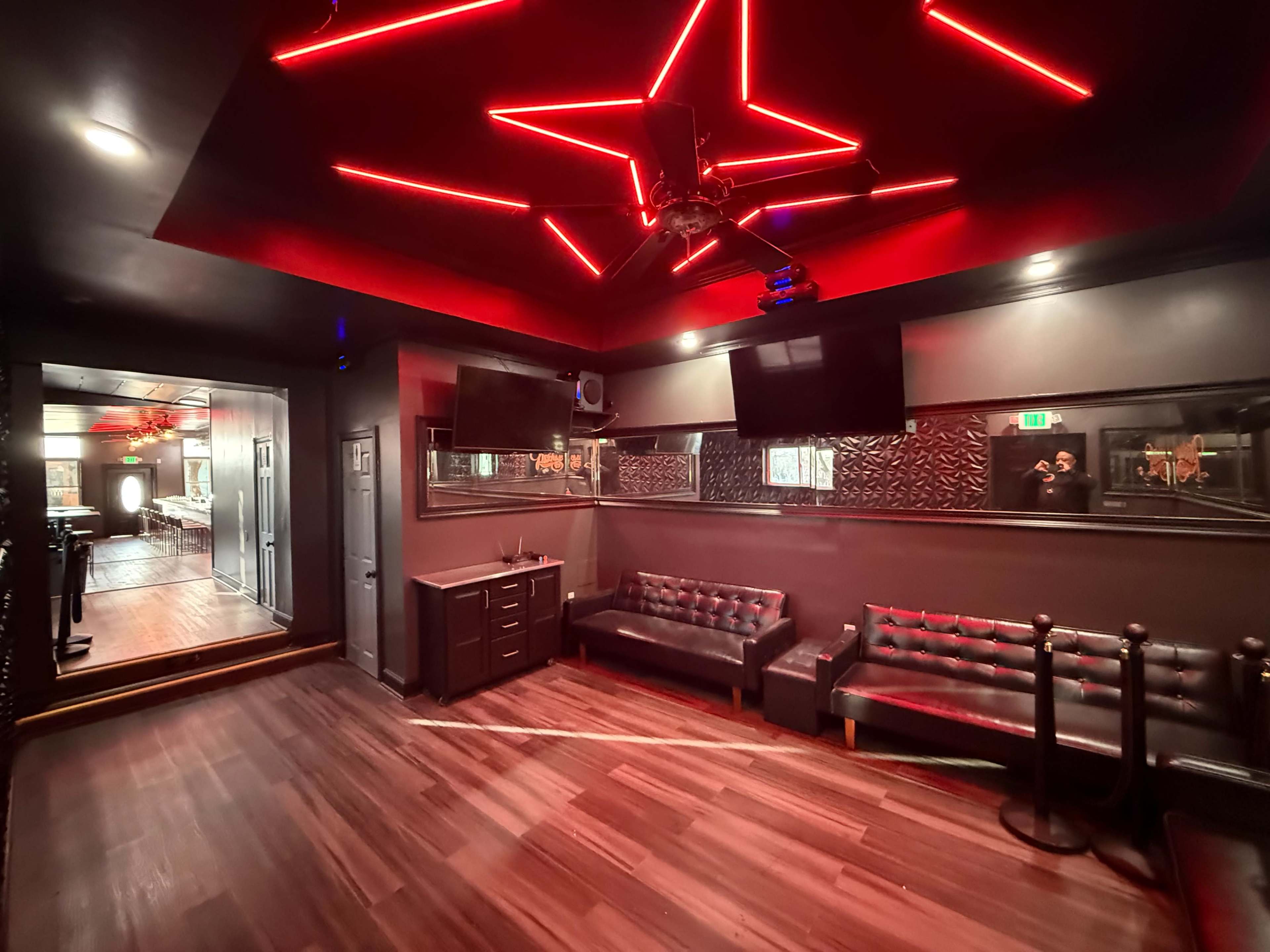 The image shows a dimly lit lounge area with black leather couches, red neon lighting, and monitors on the walls.