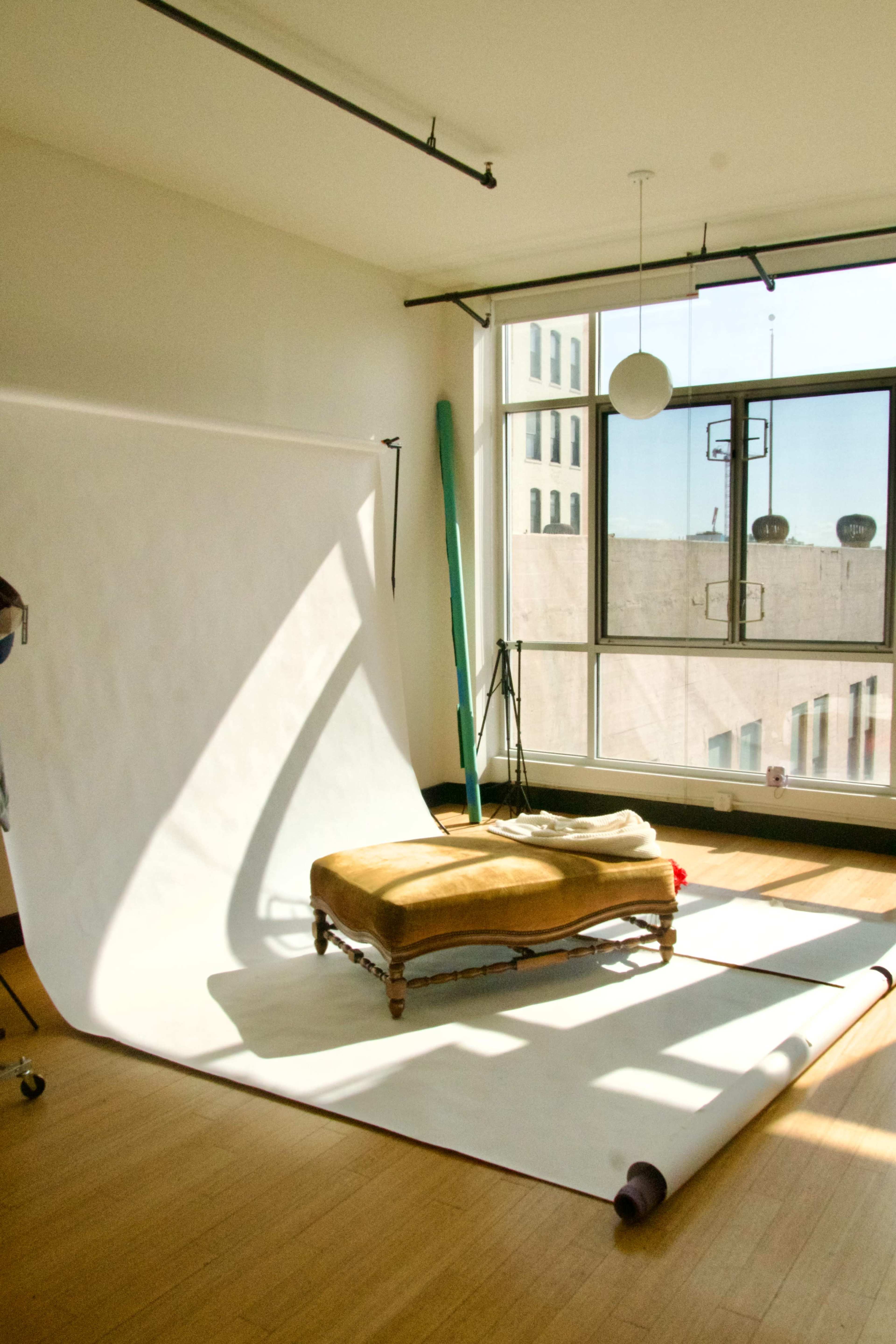 A studio space features a large white backdrop, a small upholstered sofa, and natural light streaming through a large window.