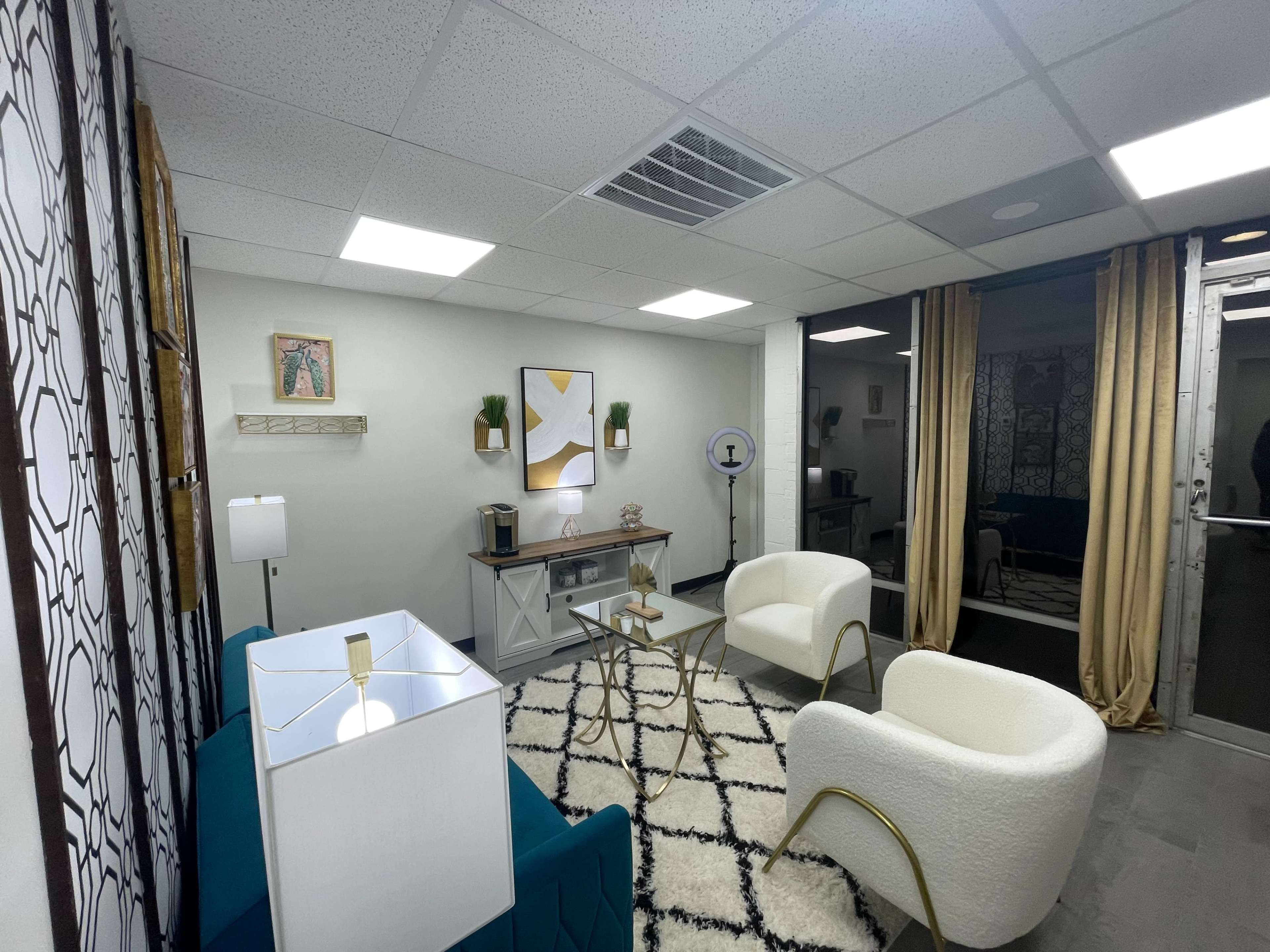 The image shows a modern waiting room with two white chairs, a glass coffee table, a blue accent sofa, and decorative wall art.