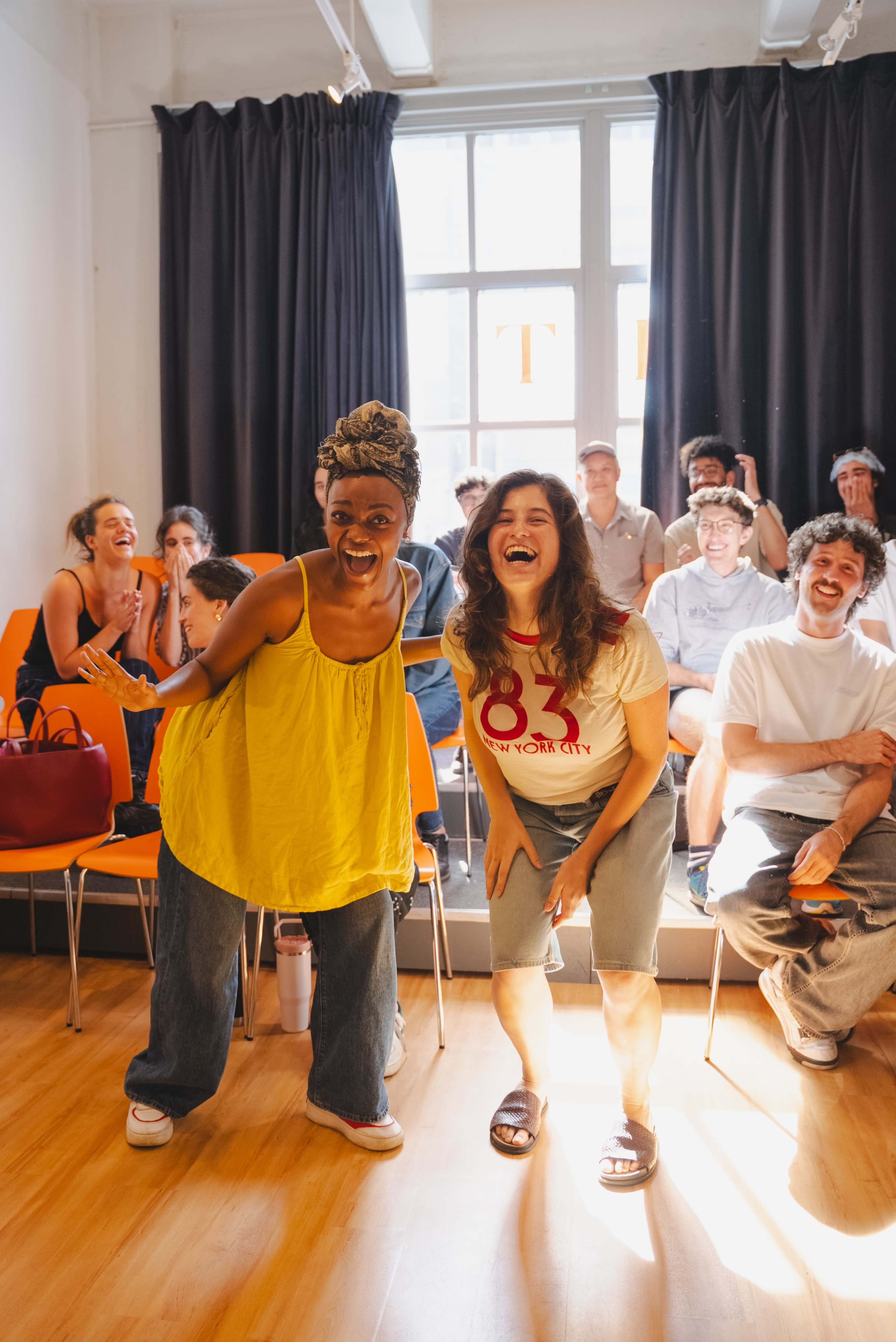 Two women in casual clothing are playfully posing in the foreground, while an audience of people is seated in the background, enjoying the moment.