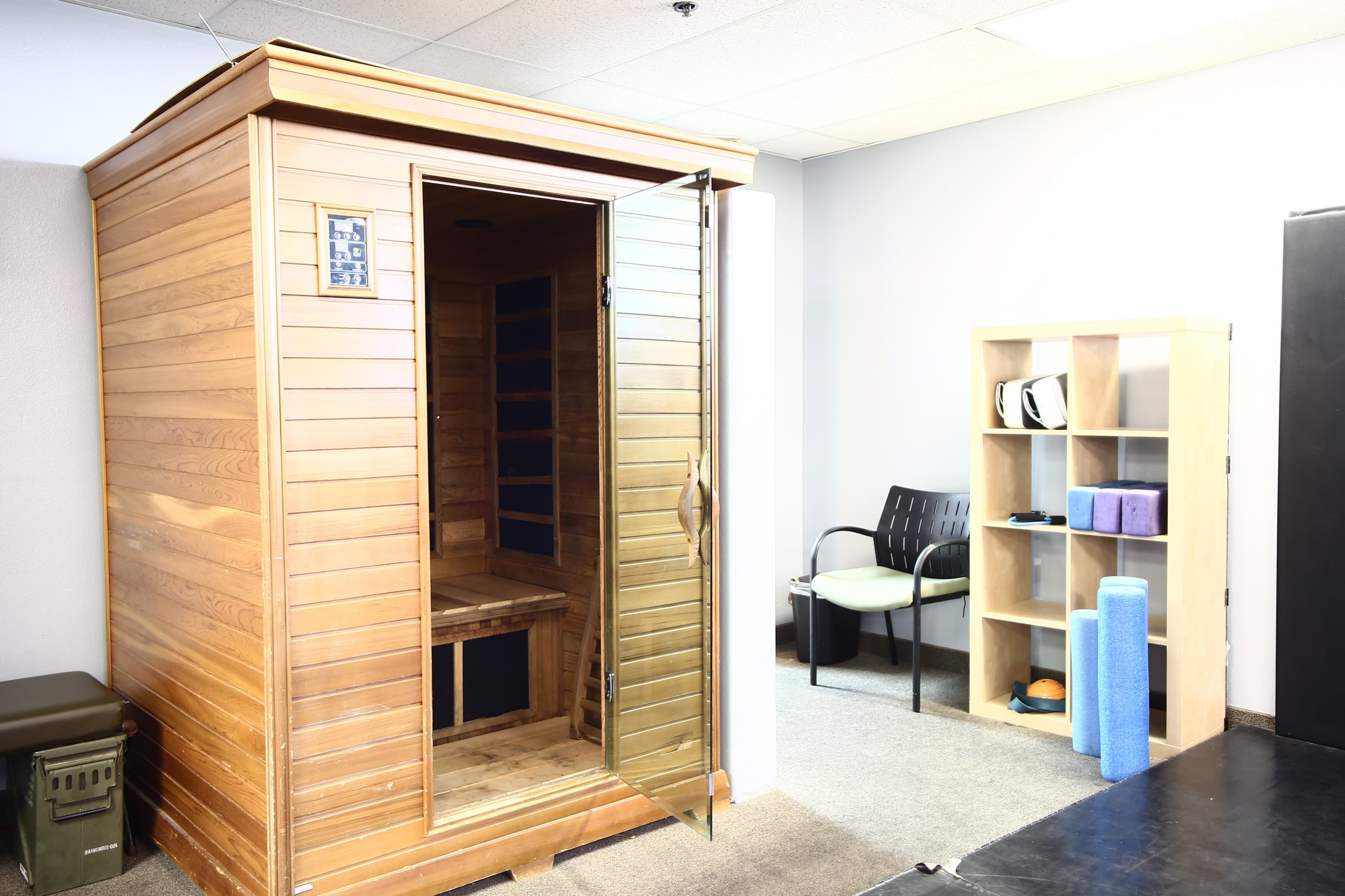 A wooden sauna with an open door, positioned beside a shelf containing various fitness equipment, in a brightly lit room.