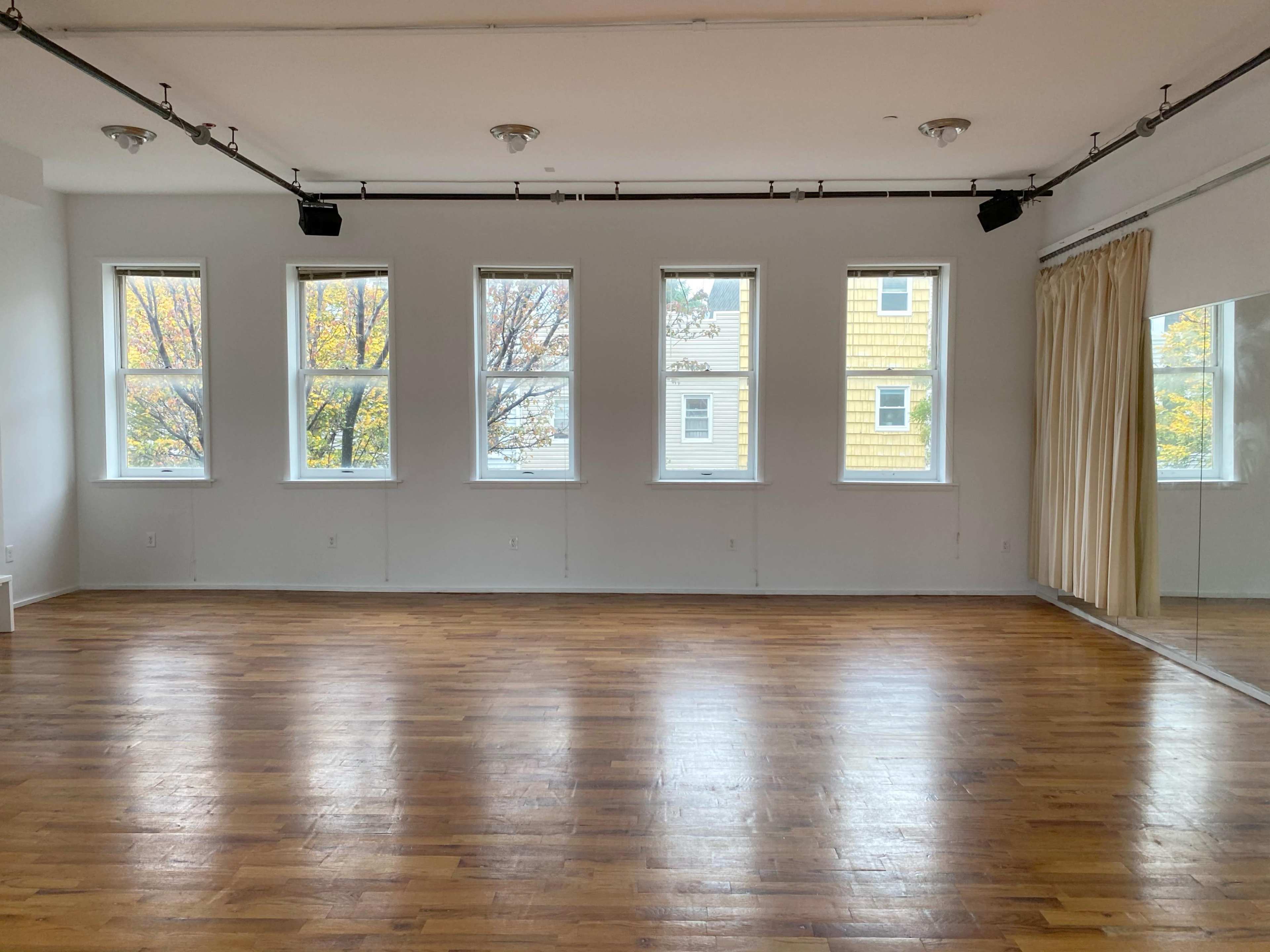 An empty wooden-floored room with large windows and a mirrored wall.