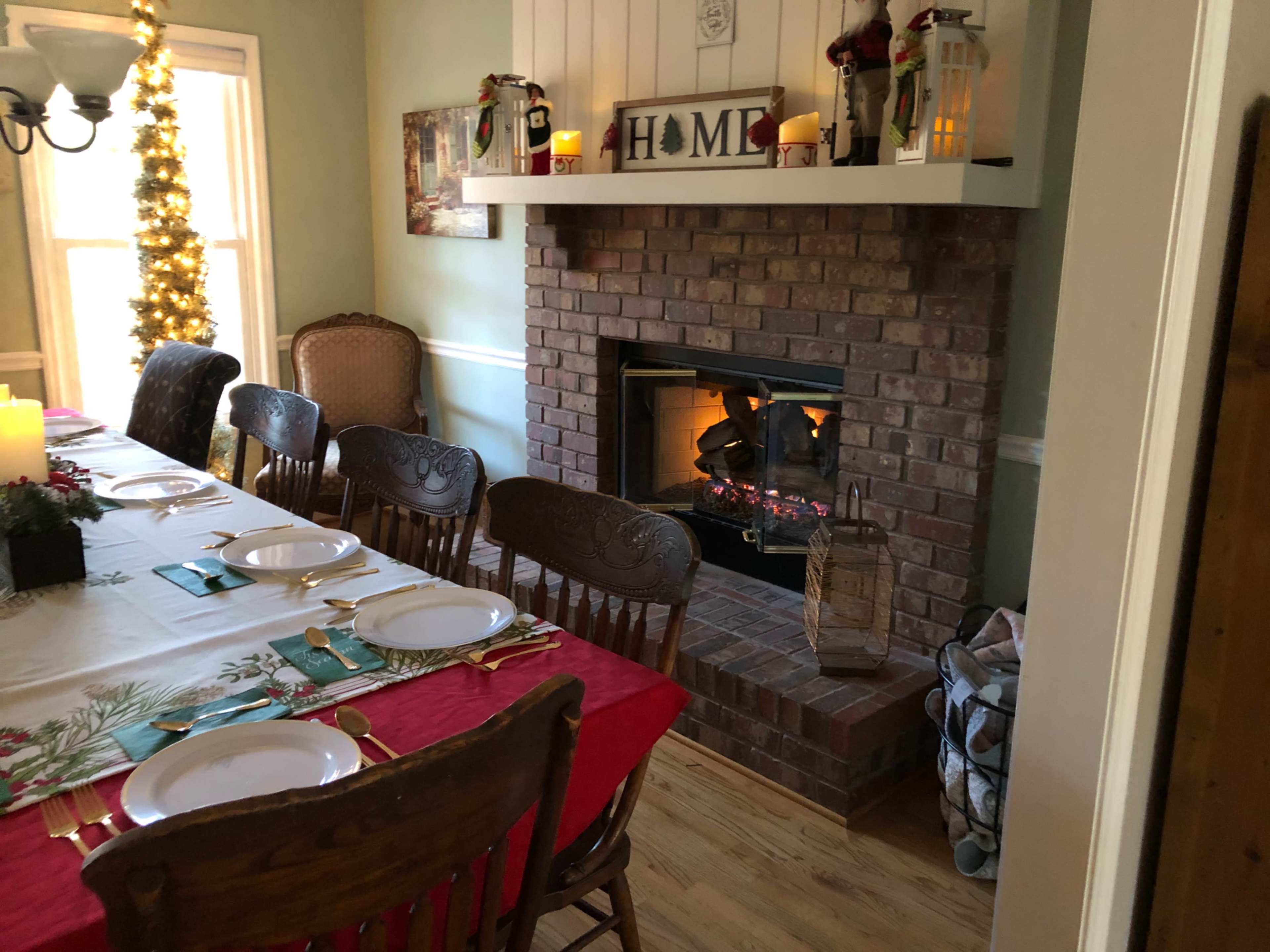 A dining area features a long table set for a meal, with a brick fireplace adorned with candles and holiday decorations.